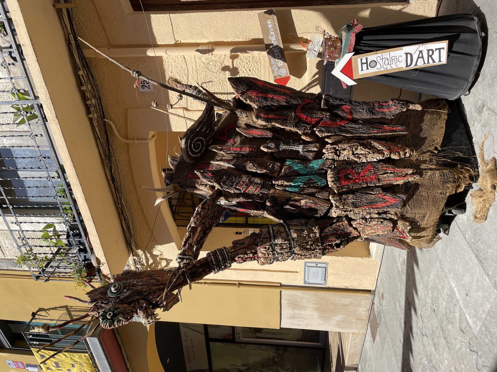 The image shows a street scene in Hostalric, Spain. An abstract, wooden sculpture stands in the foreground. The sculpture is made of stacked pieces of bark, painted with red and teal accents. A sign reading "Hostalric DART" with an arrow pointing to it is attached to the sculpture. Text including "@Sforstersteven" and "8. HOSTALRICH" is legible in the background. The buildings are painted in shades of yellow. There is a sign on the wall that reads "Aigua Potable."