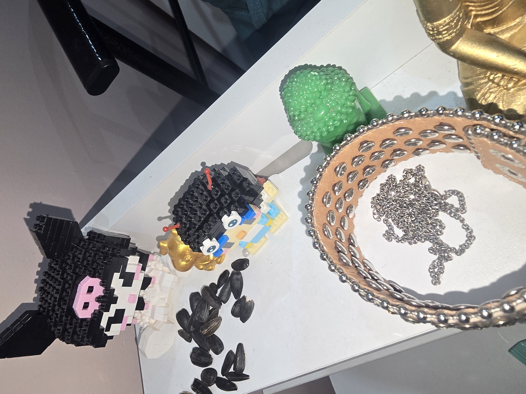 On a white surface, a collection of objects is arranged, including a black and white LEGO bust resembling the character Kuromi, a smaller LEGO figure with blue and yellow accents, a pile of sunflower seeds, a green textured figurine, and a tan leather bracelet adorned with silver studs, accompanied by a silver chain. The background consists of a pinkish-grey wall and a dark chair. The image does not contain information about the location in Ulan-Ude, Russia.