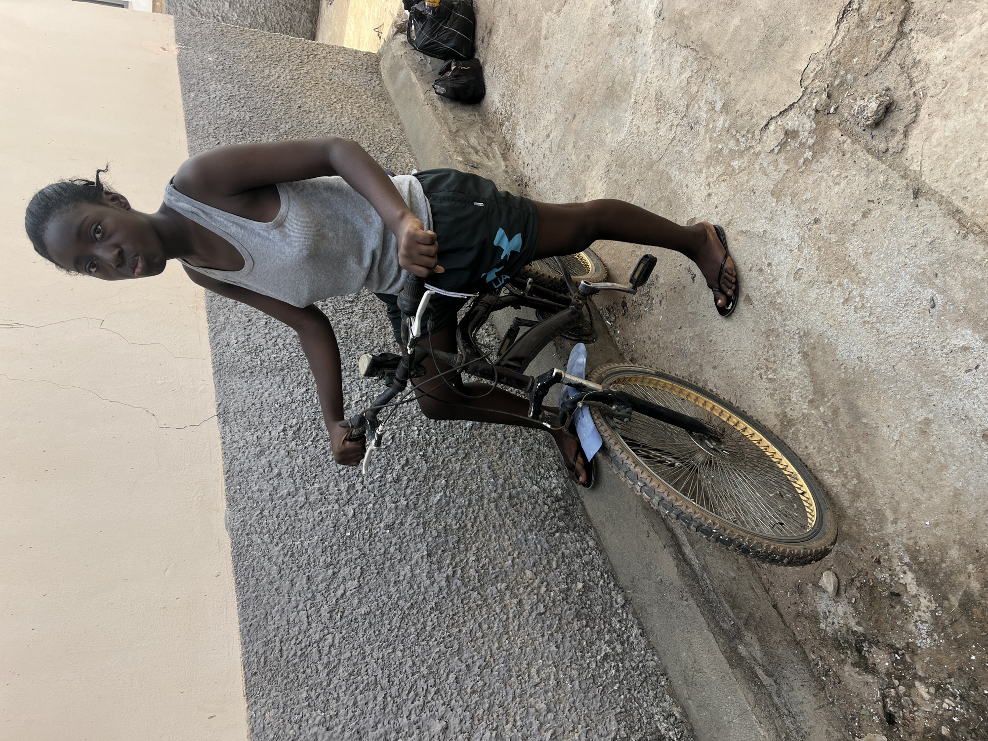 A young black woman stands astride a bicycle on a paved surface, likely a street or alleyway, in an urban setting. She is wearing a light-colored tank top and dark shorts with an "UA" logo visible on the left leg. The bicycle appears old and worn, resting against a wall. The paved surface shows signs of wear and discoloration. In the background are black bags and shoes. The location is likely Abuja, Nigeria.