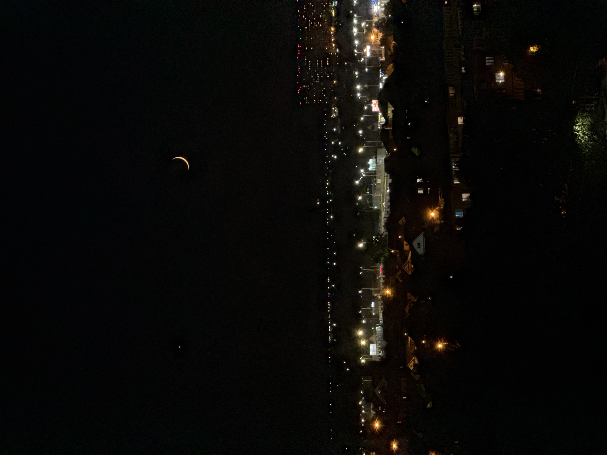 The image, taken at night, shows a crescent moon in the dark sky alongside a city skyline. The city's lights extend along a waterfront, indicating a possible river or bay. The bright lights along the water are reflected in the water. The image appears to be rotated. Based on the skyline and presence of a river, it is highly probable the city is Kazan, Russia.