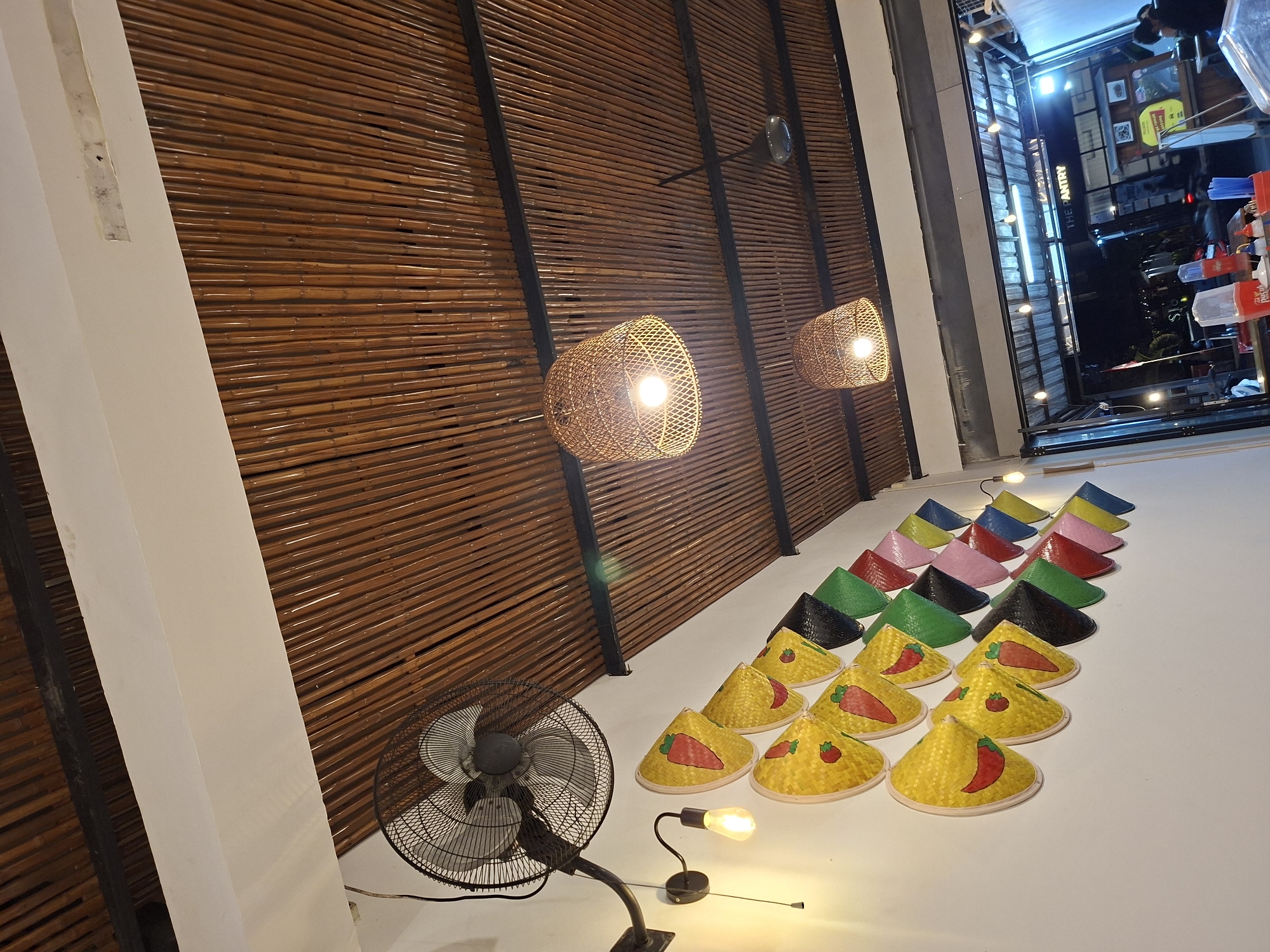 The image depicts the interior of a restaurant with a bamboo ceiling. A black oscillating fan is mounted on a white wall. Below the fan, a collection of conical hats, decorated with colorful fruit motifs, are displayed in staggered rows. The hats are yellow with red fruit, green, black, pink, and blue. Two woven pendant lamps with visible bulbs hang from the ceiling. In the background, through an open doorway, a dimly lit street scene with cars and pedestrians is visible, along with a sign that reads "THE PANTRY." The overall atmosphere suggests a casual dining establishment in Kuta, Indonesia.