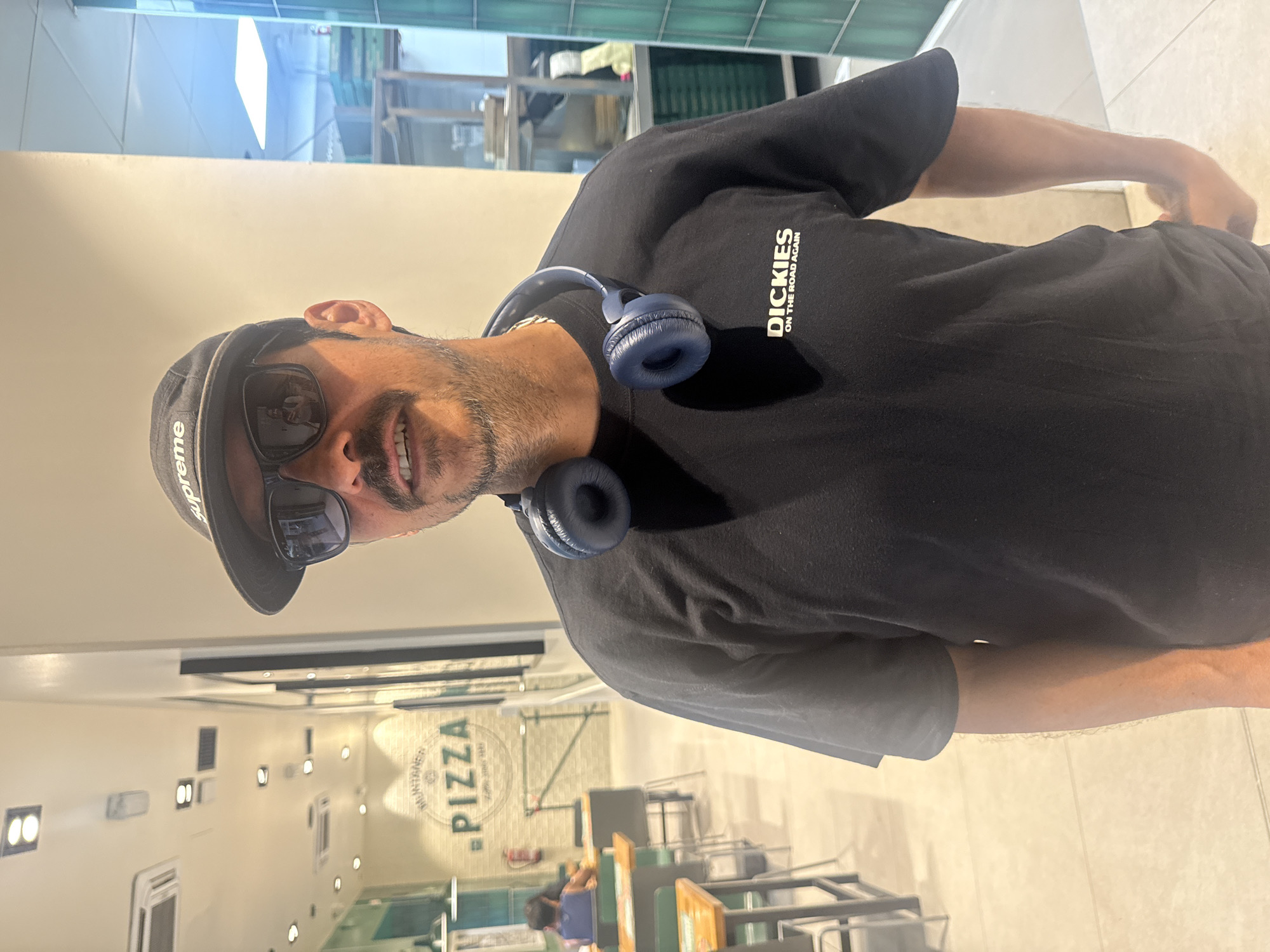 A man is standing inside a pizza restaurant. He is wearing a black t-shirt with "DICKIES ON THE ROAD AGAIN" printed on it, a black Supreme baseball cap, and sunglasses. He also has blue headphones around his neck.
The restaurant has a sign that reads "PIZZA" in large letters. There are tables and chairs visible in the background, and a person can be seen seated at one of the tables. The walls are white, with several light fixtures. The interior appears clean and modern.
It is impossible to confirm the city of Barcelona, Spain based on the details in the image.