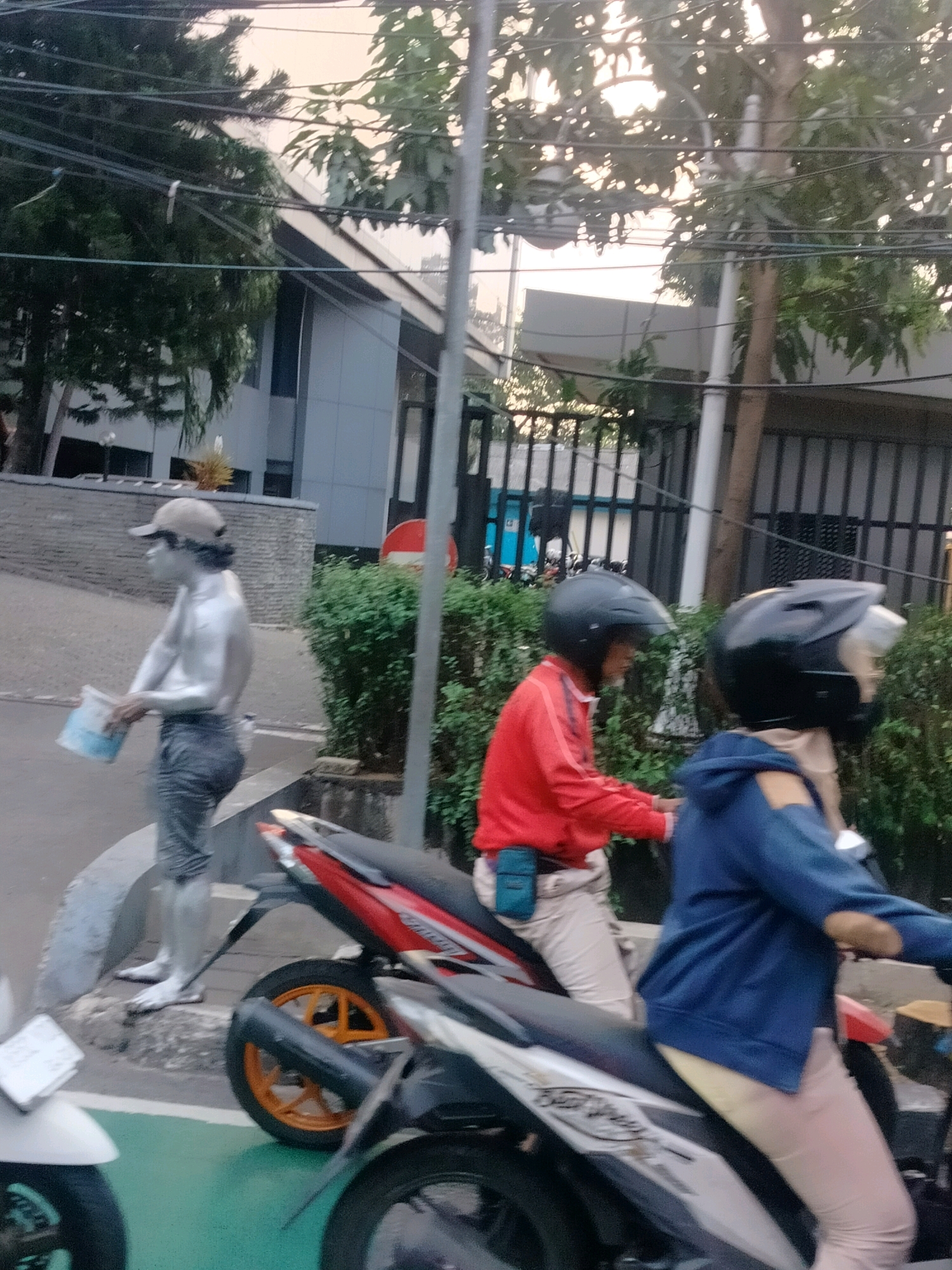 The image depicts a street scene in East Jakarta, Indonesia. In the foreground, two people are riding motorcycles. The person in the foreground right is wearing a blue hooded jacket and a black helmet, with light-colored pants. The person on the motorcycle behind is wearing a red jacket and a black helmet, with light-colored pants.
To the left, a person is standing on a curb, appearing to be covered in silver paint from the waist up, with dark pants and a beige cap. This individual is holding a white container with blue markings. Behind the people, there is dense green foliage and a fence. A modern building with a grey facade and dark windows is visible in the background, partially obscured by trees and overhead wires. The lighting suggests it is daytime, possibly late afternoon.