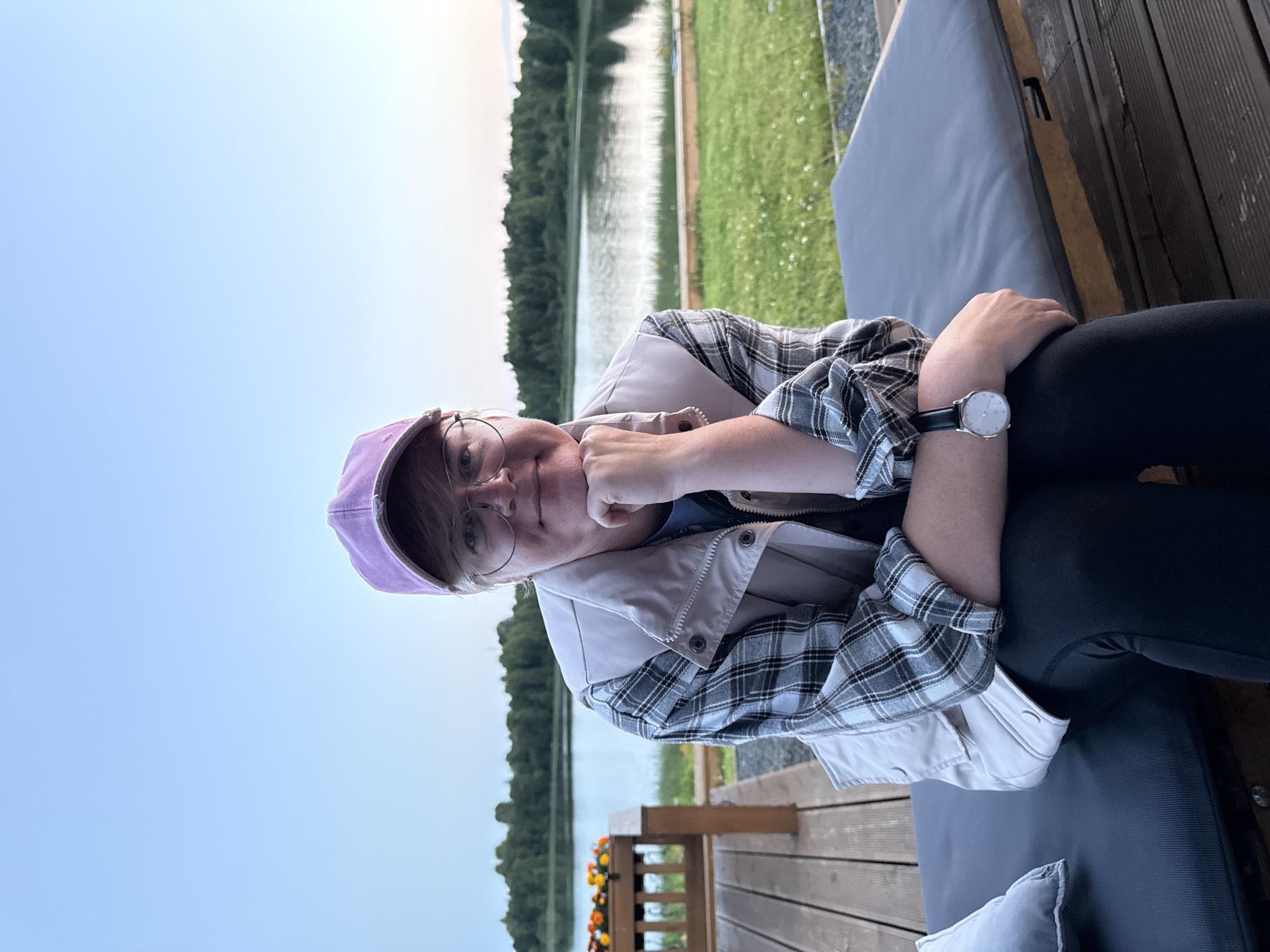 A person is sitting on a dark gray cushioned bench outdoors. The person is wearing a light-colored vest over a gray and white plaid shirt, black leggings, a purple baseball cap, and glasses. Their left hand is resting on their chin, and they are wearing a watch on their left wrist.
Behind the person, a body of water is visible, with trees lining the opposite shore. A wooden deck is in the foreground, and a railing with flower boxes is to the left of the bench. The sky is a light blue, suggesting it is either early morning or late evening. The scene appears to be a serene natural setting, possibly a park or a lakeside recreational area.
The location is identified as Pyhäjärvi, Russia, a city known for its numerous lakes and natural beauty. The image captures a moment of quiet repose in this tranquil environment.