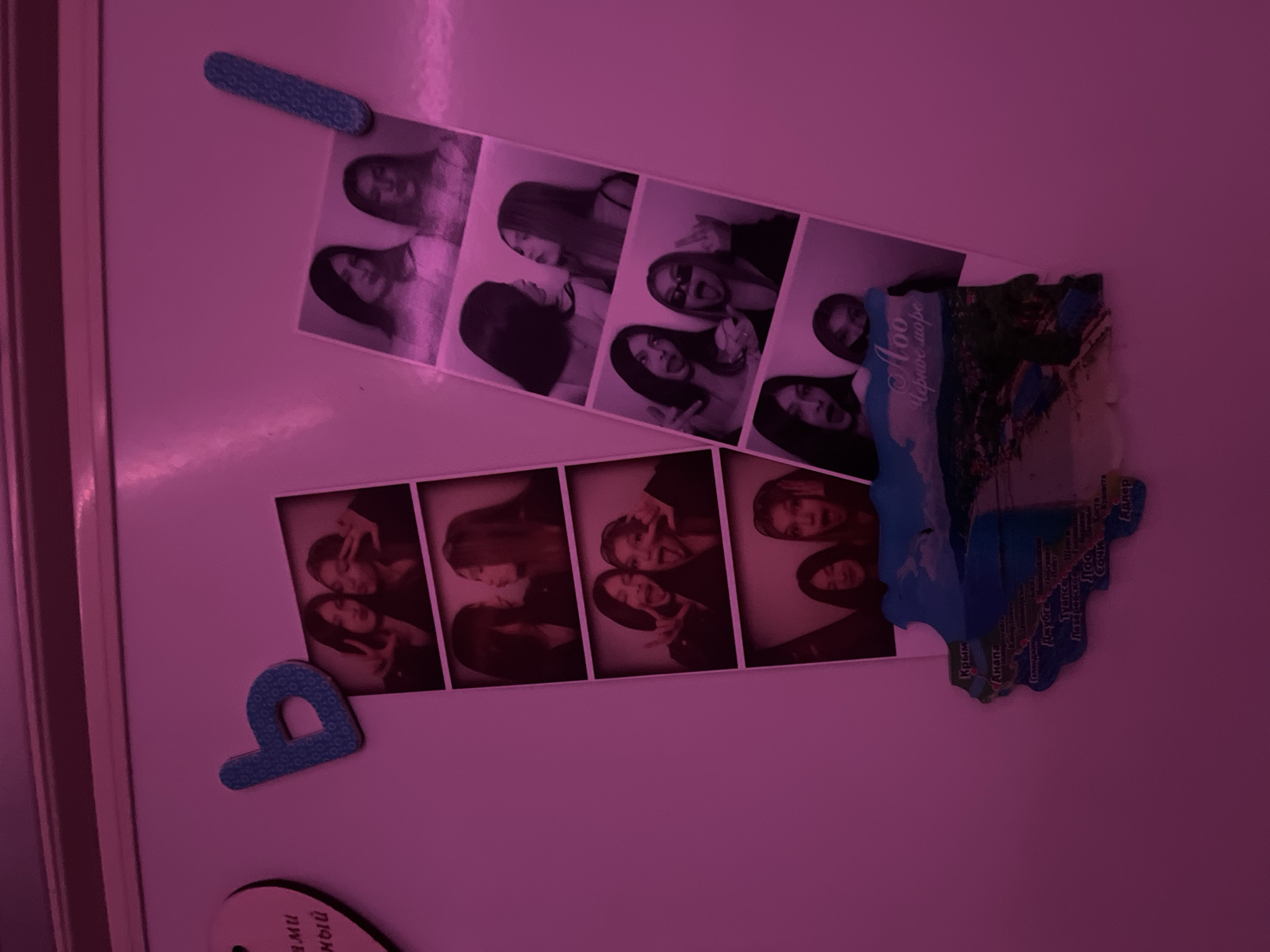The image shows a white surface, likely a refrigerator door, with several items attached to it. There are two strips of black and white photo booth style pictures of two young women. In the photos, the women are seen posing in various ways. There is also a blue magnet shaped like the letter "p", a long rectangular magnet, and a souvenir magnet that is shaped like the Crimean Peninsula. The text on the Crimean Peninsula magnet reads "Aoo Чёрное море". Some of the place names visible on the magnet include Anapa, Gelendzhik, Sochi, and Adler. The lighting of the image gives it a pink hue.