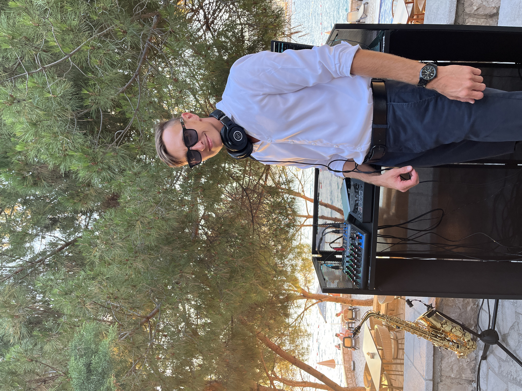 A man stands outdoors, with a DJ setup and a saxophone beside him, in front of a body of water. The man is wearing a white dress shirt, dark pants, sunglasses, and headphones around his neck. He is smiling and looking at the camera. The DJ setup includes a mixer and what appears to be a laptop. The saxophone is gold-colored and on a stand. In the background, there are trees with green foliage, a beach with lounge chairs and umbrellas, and several people relaxing. The lighting suggests it is late afternoon or early evening, with the sun casting a warm glow. The location is identified as Pečurice, Montenegro.