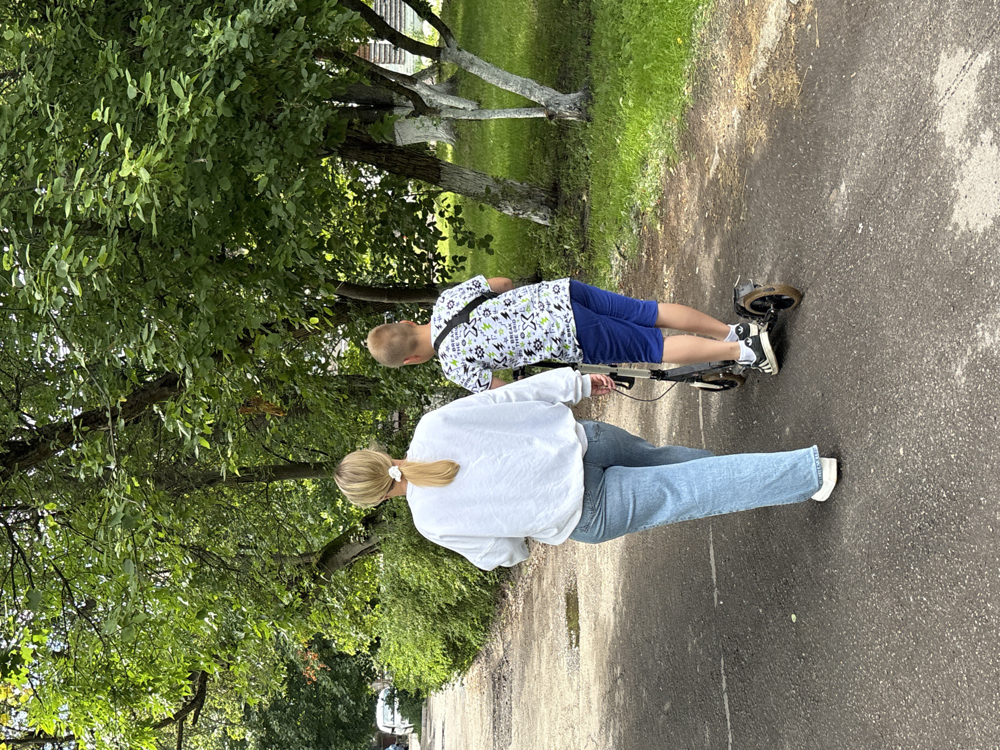 The image shows two people on a paved path outdoors. In the foreground, a woman with blonde hair pulled back in a ponytail is wearing a white sweatshirt and light blue jeans. She is walking and holding the handlebars of a scooter. Behind her, a boy is riding on the same scooter. He has short blonde hair and is wearing a white t-shirt with a black and green pattern and blue shorts. He is wearing black and white sneakers. The scooter has black wheels and a black frame.
The scene is set in a park-like area with lush green trees and grass on the left side of the path. Some of the trees have white paint on their trunks. The path appears to be made of asphalt and has some dirt and dry grass patches alongside it. In the background, there is more greenery, and a hint of a white building can be seen through the trees. The lighting suggests it is daytime, and the weather appears to be pleasant, suitable for outdoor activities. There is no visible text in the image. The overall mood is relaxed and active, with the two individuals enjoying a ride together. The location context is Saint Petersburg, Russia.