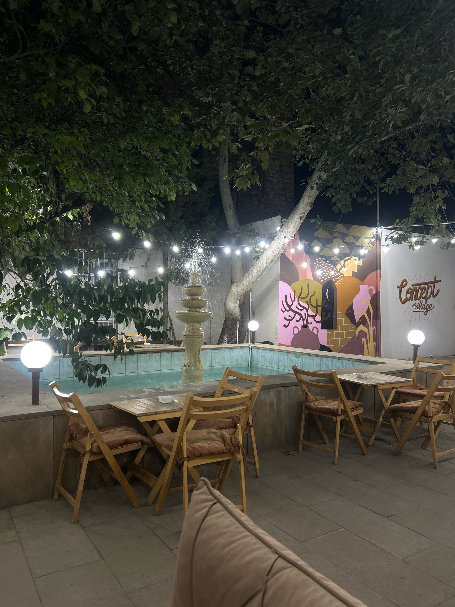 A fountain with multiple tiers is illuminated in the center of a courtyard with light strings overhead. To the right of the fountain, a mural depicts abstract figures and jugs, and the words "Concept Village" are written in script. A pool surrounds the fountain, and seating areas with wooden tables and chairs are visible. The scene is lit by the fountain's glow and string lights. The image is likely taken in Baku, Azerbaijan.