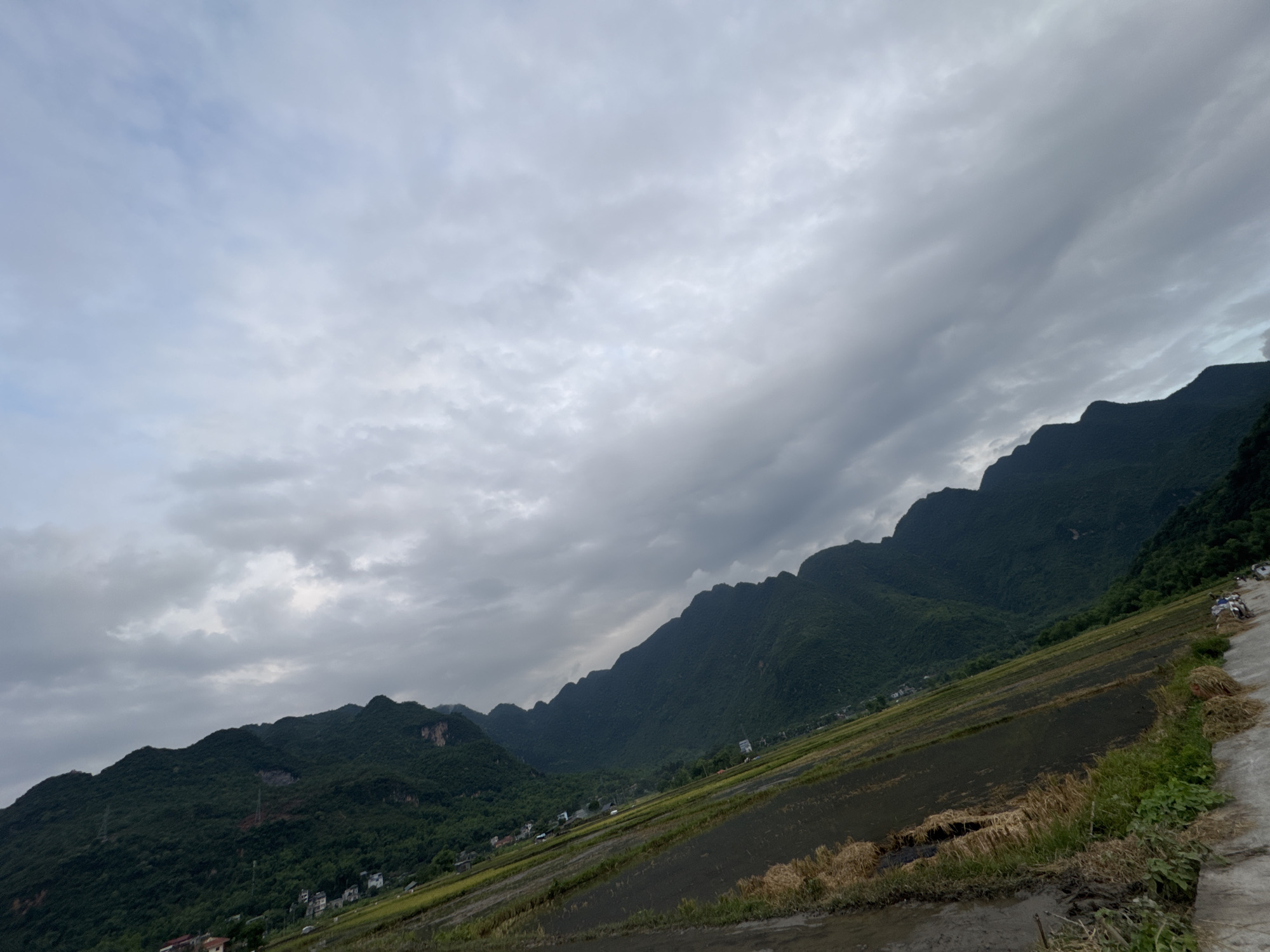 A wide-angle shot captures a rural landscape in Chiềng Châu, Vietnam, under a cloudy sky. The foreground features a sloped, muddy field with some harvested straw, indicative of agricultural activity. A concrete path runs along the right edge of the frame, with a few motorcycles parked near it. In the middle ground, terraced rice paddies stretch across the hillside, interspersed with small clusters of houses. The background is dominated by a range of lush, green mountains with jagged peaks, their slopes covered in dense vegetation. The overall lighting suggests it might be overcast or late in the day.
