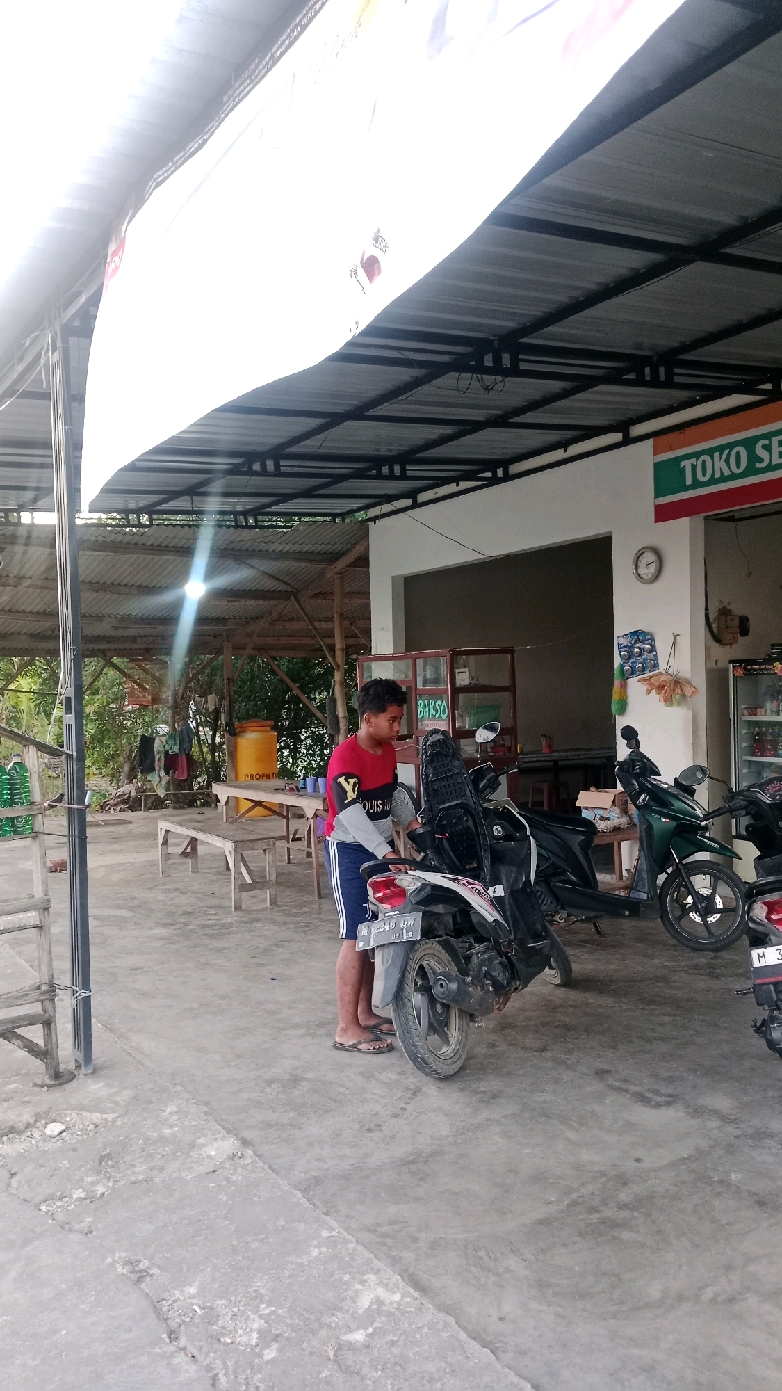 In Dempo Barat, Indonesia, a young boy in a red and gray shirt with blue shorts stands next to a white and black motorcycle under a corrugated metal roof. He appears to be adjusting something on the motorcycle. To his right, another motorcycle, dark green, is parked. In the background, a store with a sign reading "TOKO SE" and "BAKSO" is visible. The area is paved with concrete. To the left of the boy, there are wooden tables and benches, a yellow water tank, and some hanging laundry. The overall environment suggests an outdoor seating area attached to a small shop.
