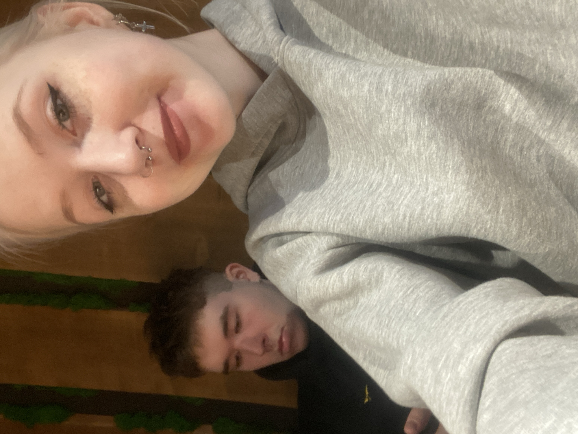A selfie photograph shows a young blonde woman and a young man in an indoor setting. The woman has fair skin, blonde hair, and is wearing a grey hoodie. She has piercings in her nose and ear, and her eyes are lined with black eyeliner. The man is behind the woman, and he appears to be either sleeping or resting his head on her. The background is blurry, but there is a wooden panel with green foliage attached to it. There is not sufficient information to determine the location in the city of Izhevsk, Russia.