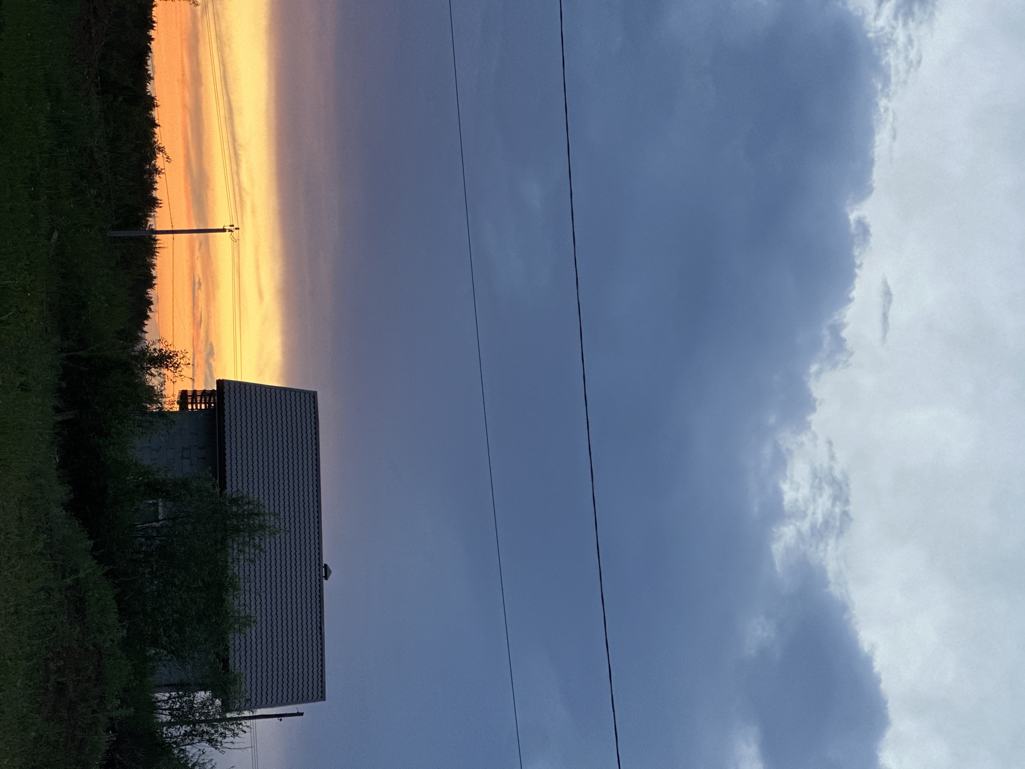 The image shows a house in Прикамский, Russia, silhouetted against a sunset. The house has a grey roof with dark tiles. There are trees in front of the house. The sunset is a bright orange color fading into a grey sky. There are power lines crossing the sky. The left side of the sky is cloudy.