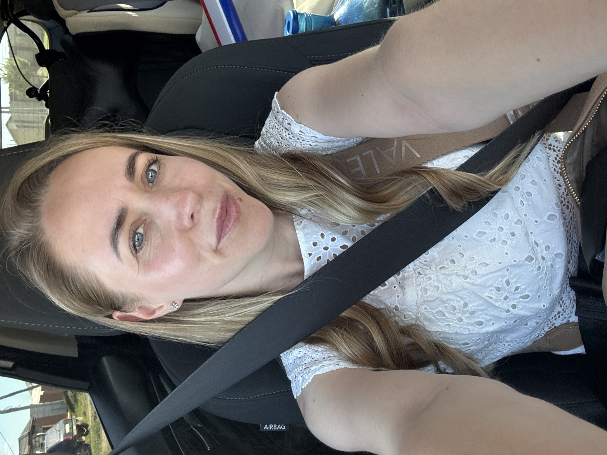A woman with blonde hair and blue eyes is seated in a car wearing a white lace shirt. She is wearing a seatbelt. A beige-colored bag is visible near her shoulder. The location appears to be Chaikovsky, Russia based on a building visible through the car window in the original image. A truck and some greenery are also seen outside the car window.