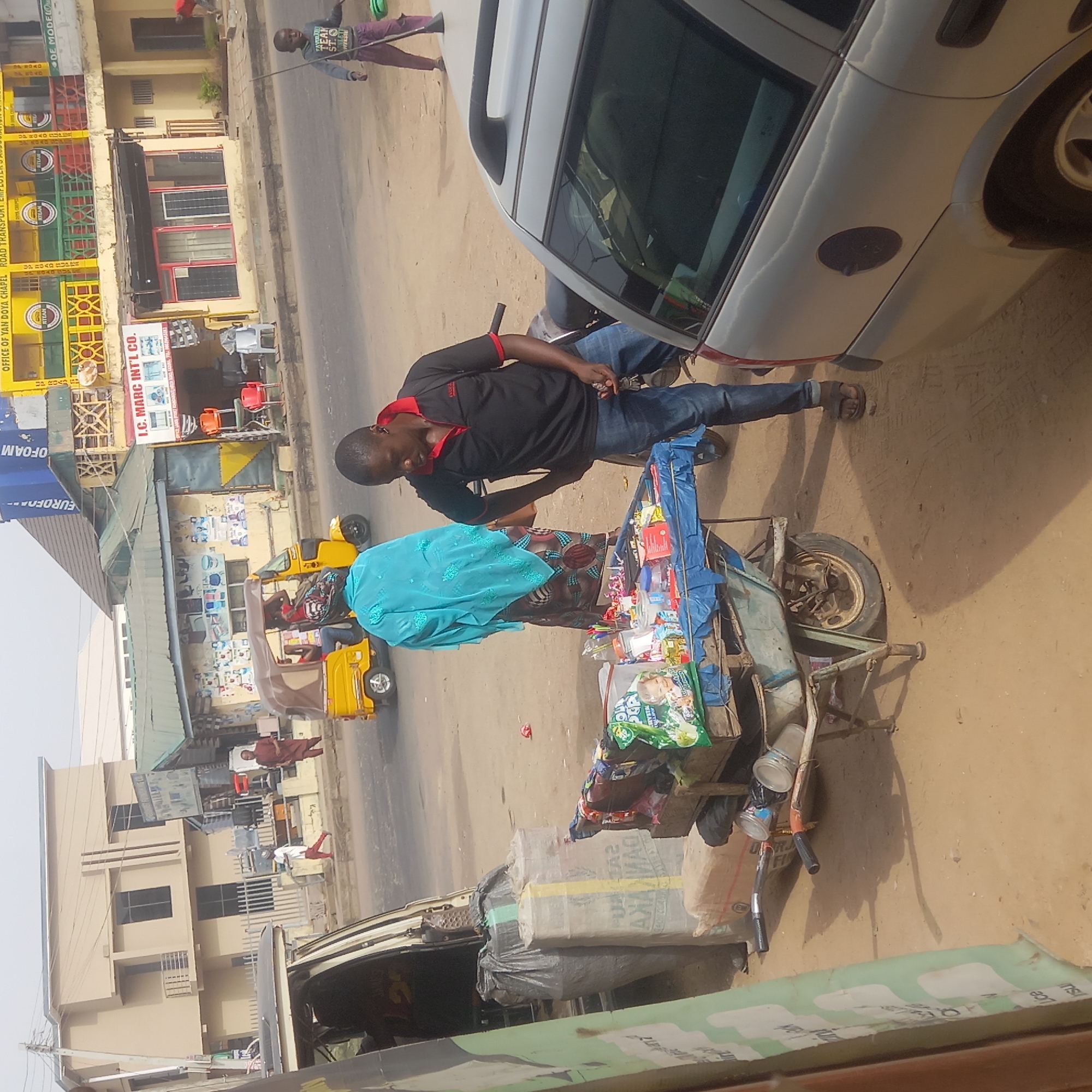 The scene is a street in what appears to be Dadi Riba, Nigeria. A man in a black shirt with red trim and blue jeans stands next to a gray car. Beside him is a woman in a colorful dress and a blue shawl, tending to a three-wheeled cart piled with goods for sale, including packaged snacks and a bag of baby formula. A yellow tricycle taxi (keke) is visible in the street behind them. Buildings with commercial signage ("I.C. Marc Int'l Co." and "Office of Yan Doya Chapel Road Transport") line the street. Other people can be seen walking in the background. The ground appears to be dirt and sand.