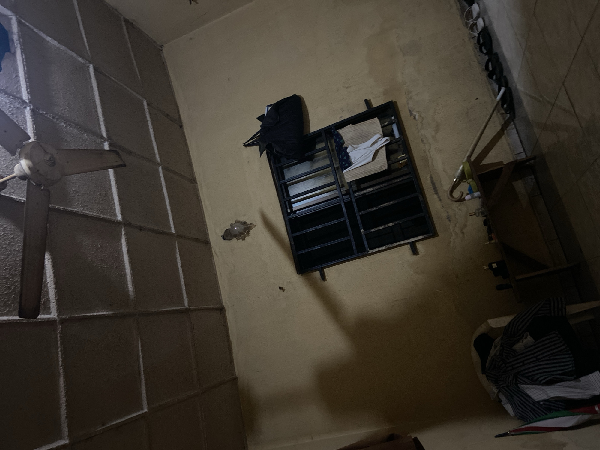 The image depicts an interior scene, likely a room, dimly lit and appearing somewhat cluttered. The ceiling features a textured surface with a grid pattern, and a ceiling fan with light-colored blades is visible. The walls are painted a pale yellow and show signs of age and wear.
A barred window is a prominent feature, with clothing items, including what appears to be a white tank top and patterned fabric, hung on the bars. A dark-colored bag is also hanging on or near the window frame. On the wall above the window, there is a small, indistinct object, possibly a light fixture or decoration. A large shadow, suggestive of a person, is cast on the wall to the left of the window.
In the foreground, on the right side of the image, a row of flip-flops or sandals are lined up on the tiled floor. To the left, a plastic chair is covered with folded clothing, including a striped shirt. A small wooden table or shelf is positioned in the center of the room, with some items on it, including what looks like a bottle and a mop with a yellow handle.
The overall impression is that of a modest living space. The specific location in Jalingo, Nigeria, cannot be determined from the visual information provided in the image.