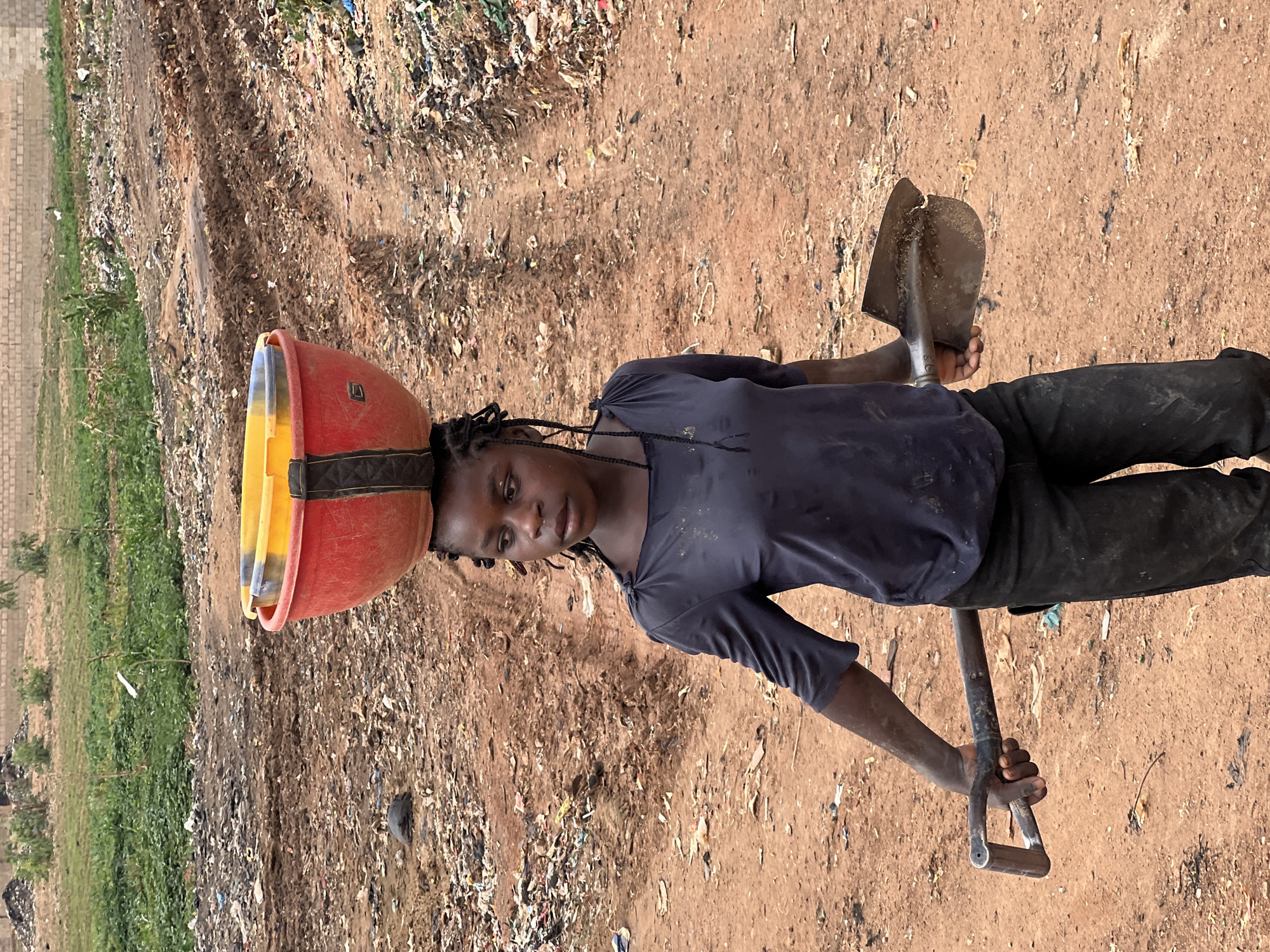 A young woman stands in a waste dump in Du, Nigeria. She balances a red and yellow bowl on her head, and holds a shovel in her hands. The ground is covered in waste, with trash piled high in the background. The woman is wearing a dark shirt and pants.