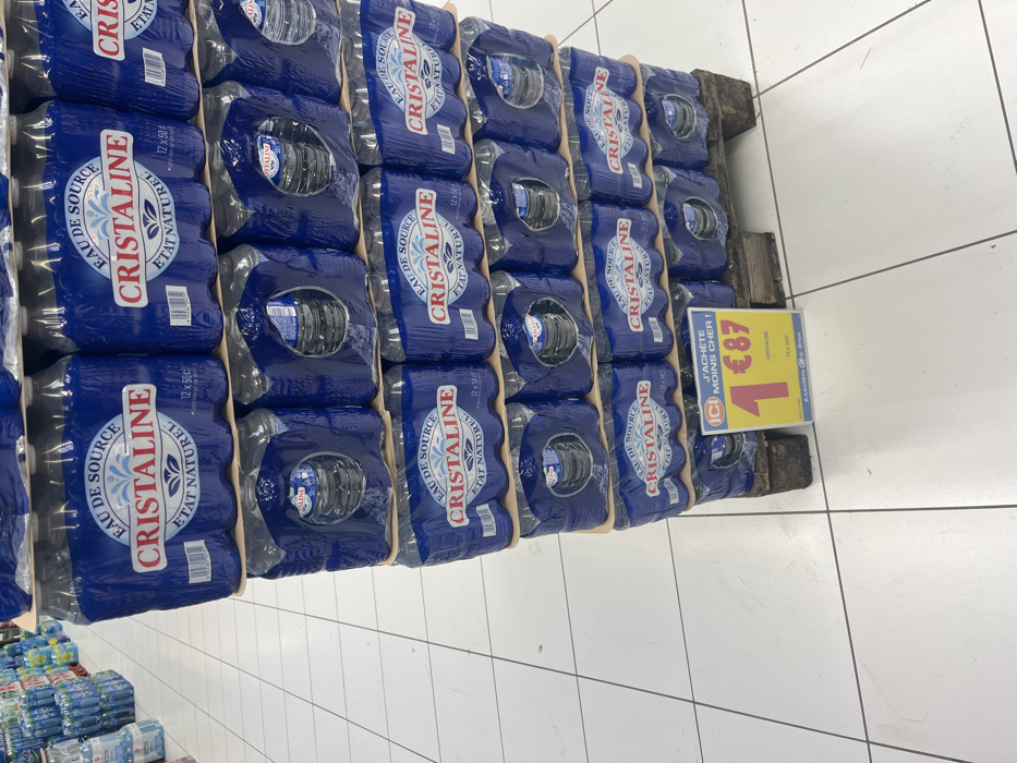 A stack of six packs of Cristaline brand bottled water is on a wooden pallet in a store in Saint-Brice-Courcelles, France. The bottles are arranged in six packs, with each pack containing six bottles. Each bottle has a blue label with the words "Cristaline Eau de Source Etat Naturel" on it, and the pallet is made of wood. A yellow sign in front of the pallet reads "J'achete moins cher!" with a number 1 and the price "€87" displayed prominently. The store floor is tiled in white and grey. The environment is clean and well-lit. There is no other person or object visible in the image.