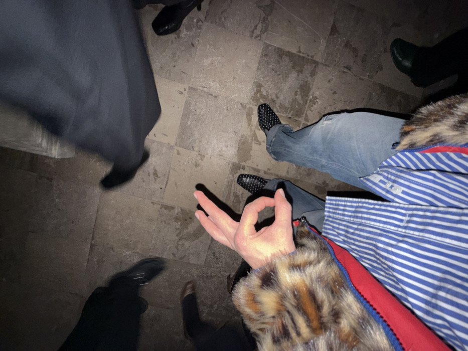 The photograph depicts a variety of people's lower extremities. One person displays the "circle game" hand gesture. Various footwear is visible, including black dress shoes and textured black flats. The floor is made of tile and has a dark, shadowy quality. A person wears blue jeans, a blue and white striped shirt, and a faux fur-trimmed red jacket. Another wears a long black dress.