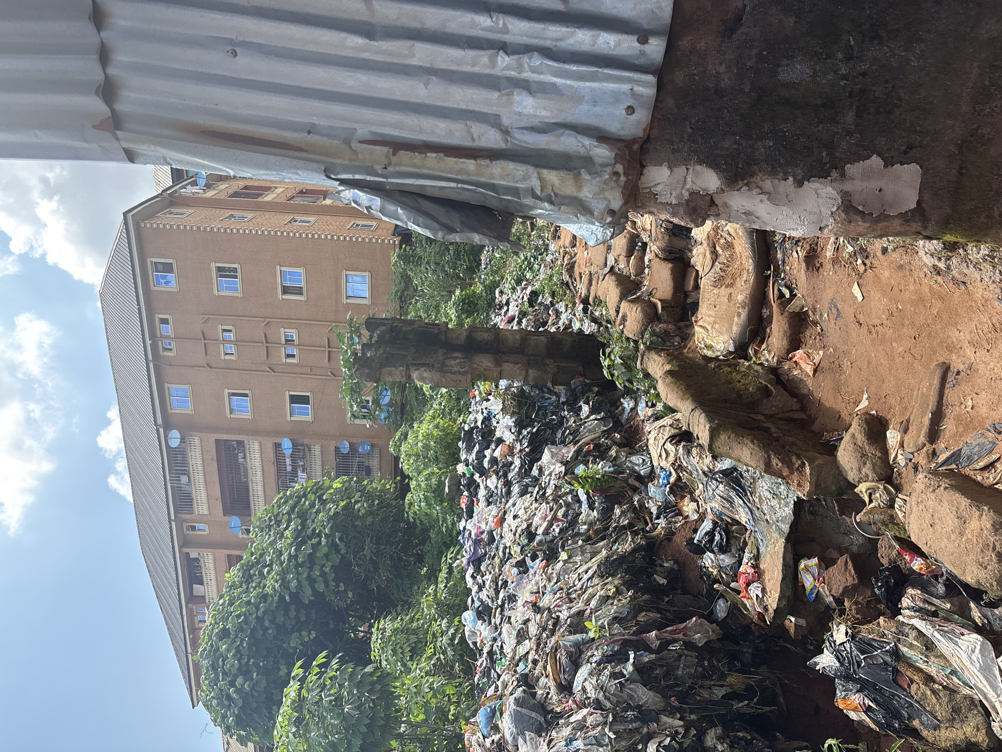 In Nkpor, Nigeria, this image depicts a large accumulation of refuse piled high against a backdrop of lush green trees and a modern apartment building. The refuse consists primarily of plastic bags and other disposable waste, suggesting a lack of proper waste management. The apartment building, with its numerous windows and satellite dishes, stands in stark contrast to the environmental degradation. A partially constructed or decaying structure made of stone and corrugated metal occupies the foreground, further emphasizing the disarray. The ground is a mixture of red earth, rocks, and scattered debris, indicating a potentially unsanitary environment. The scene appears to be an outdoor area on a bright, possibly humid day, with a partly cloudy sky overhead.