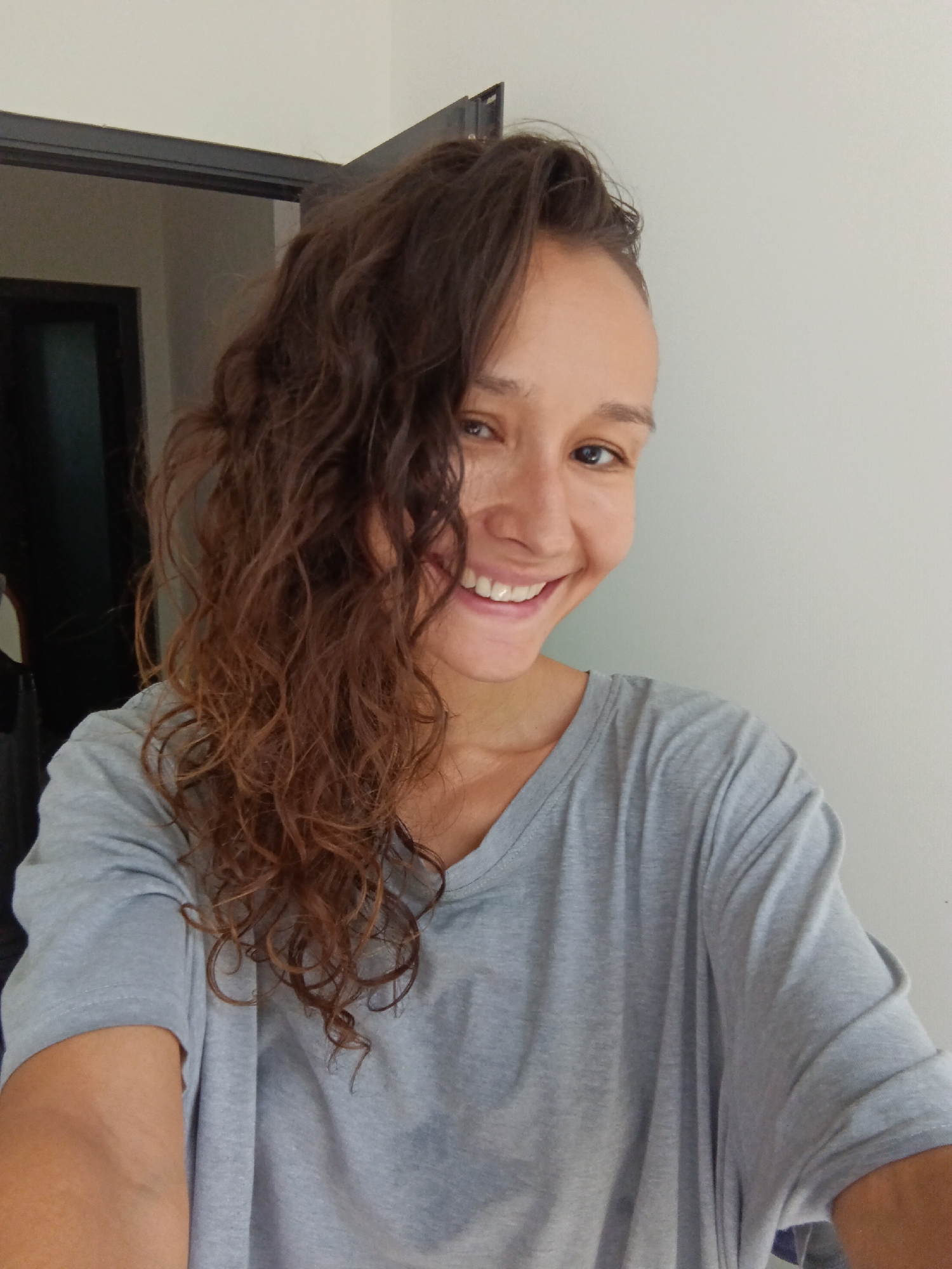 A young woman with curly brown hair and light brown skin is smiling at the camera. She is wearing a gray t-shirt and is standing in front of a white wall. The background shows a doorway with a dark interior. The lighting suggests an indoor setting with natural light coming from the right. The image is a selfie, taken from a slightly high angle. There is no indication of the city of Mũi Né, Vietnam.