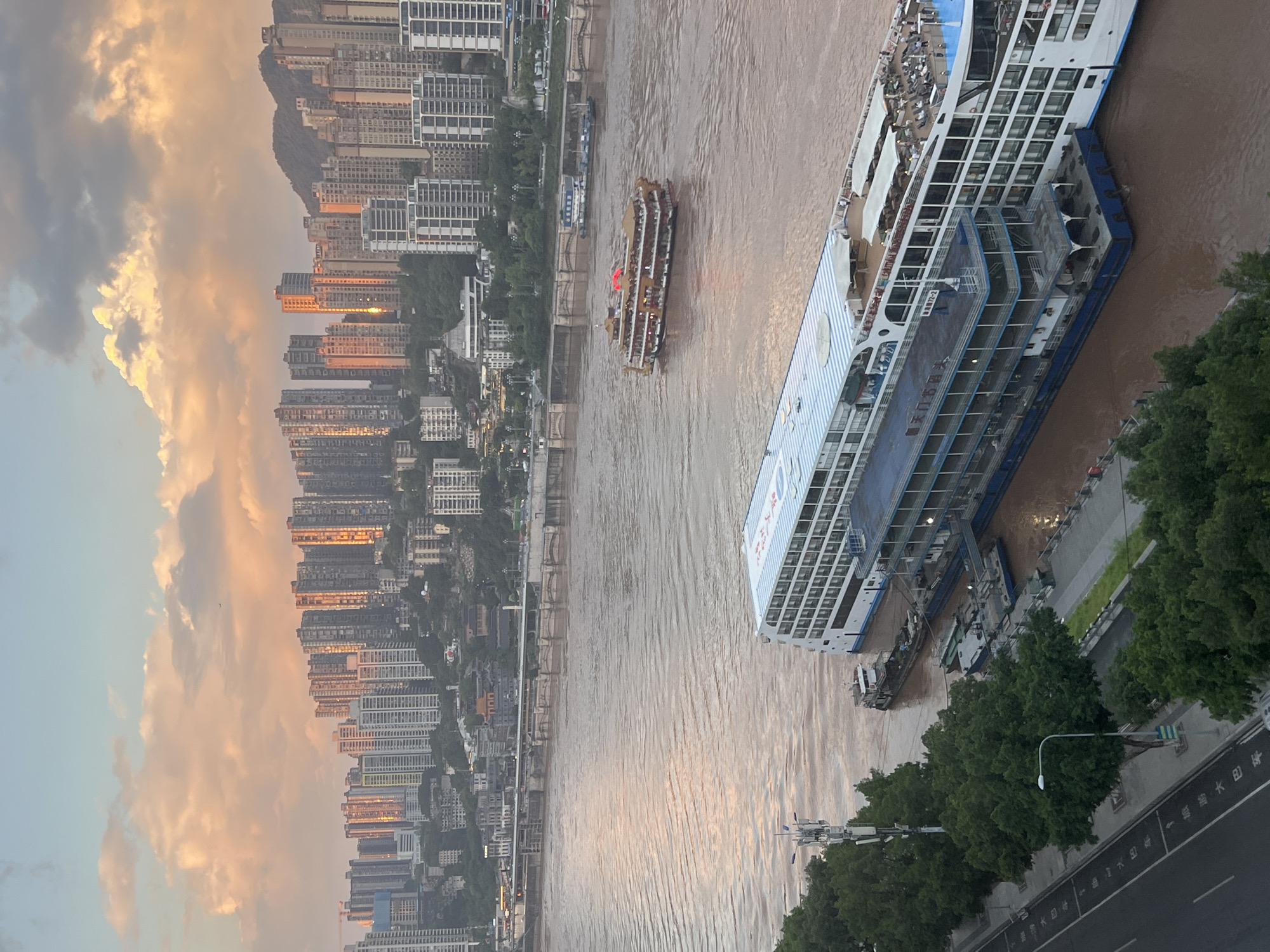The image depicts a riverfront scene in Yuzhong District, China, likely at sunset given the warm lighting on the buildings and clouds. A wide, muddy river occupies the foreground and middle ground. Several large cruise ships are moored along the riverbank. The largest of these ships, a white vessel with multiple decks, is docked closest to the viewer. Another, more ornate, golden-brown ship with red accents is actively navigating the river. Behind the river, a dense urban landscape rises up a hillside, characterized by numerous tall, modern buildings. The sky is a blend of soft blues and oranges, with scattered clouds catching the sunset light. Trees line the riverbank in the foreground, and a road with lane markings is visible to the left of the frame. The scene appears to be a busy port area integrated with a large city.