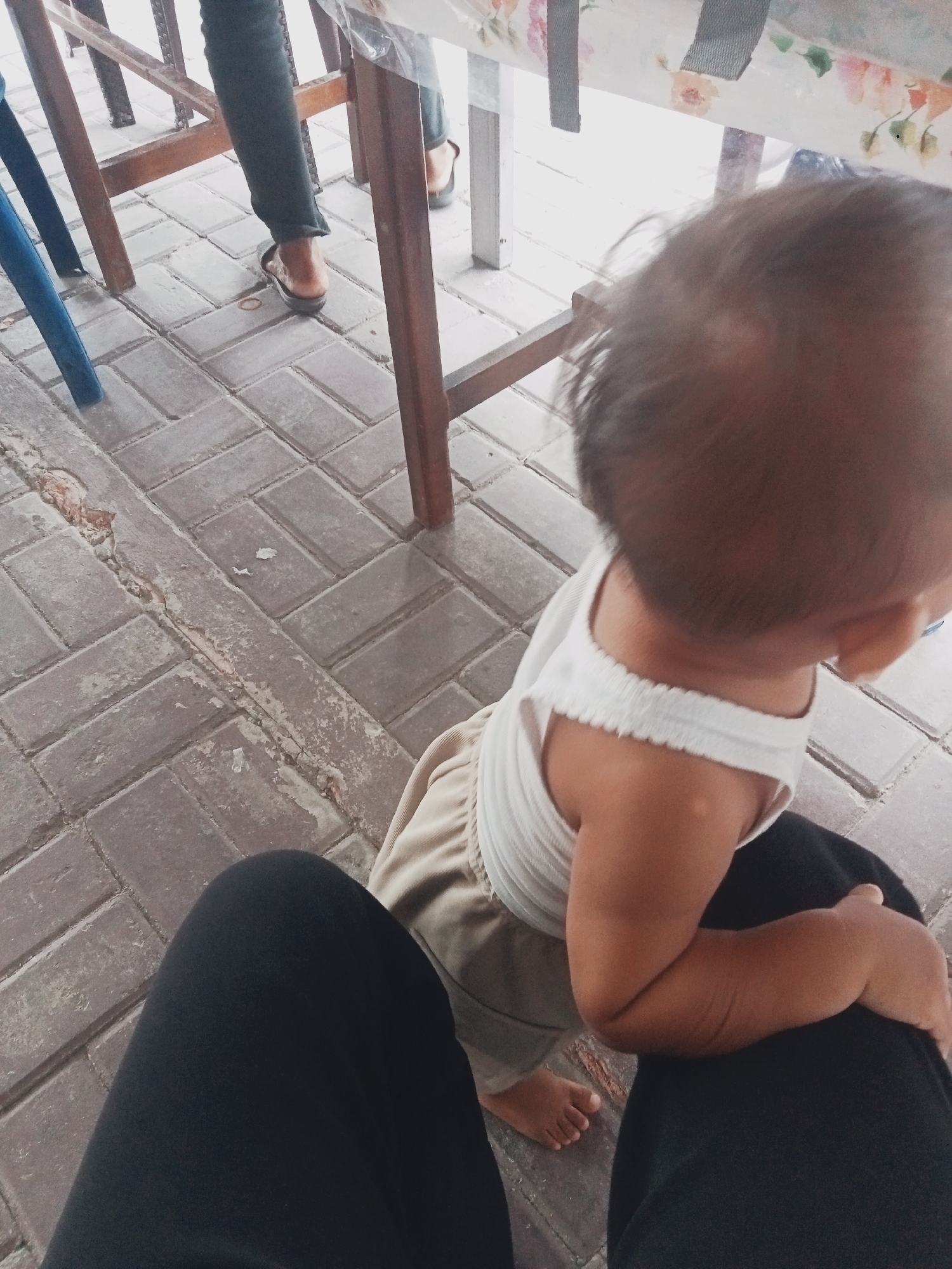 The image depicts a low-angle, close-up view of a toddler sitting on someone's lap. The toddler is facing away from the camera, with their head and upper back visible. They are wearing a white ribbed tank top and light-colored pants. The person on whose lap the toddler is sitting is wearing black pants. In the background, the legs and feet of another individual wearing dark pants and sandals are visible, standing near a wooden table and chairs. The ground is paved with rectangular bricks. The scene appears to be outdoors, possibly in a public area or a casual dining setting. The lighting suggests it is daytime. The overall impression is a candid moment captured from a personal perspective. The location of Balik Alam, Indonesia, is not evident from the visual information in the image.