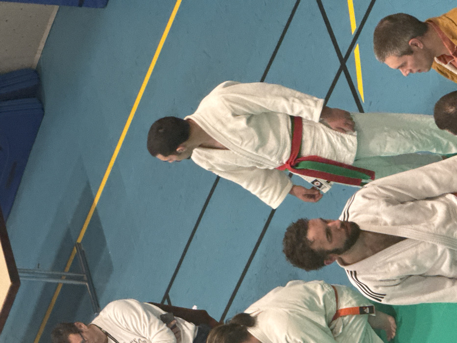 The scene is set indoors on a blue sports mat with yellow and black boundary lines, consistent with a martial arts facility in Brumath, France.
Five individuals are visible. In the foreground, a person is partially visible, kneeling or sitting on the mat, wearing a white judogi secured with an orange belt. Bare feet are present. To their right, a male with dark, curly hair and a beard stands, wearing a white judogi with three black stripes on each shoulder. He faces generally to the right, looking downwards. Behind him, another male stands, wearing a white judogi with a prominent red and green striped belt. He is looking to the left. In the background, on the far left, a male with glasses and dark hair sits on a dark chair, wearing a white long-sleeved top with partial text "FSC" visible on the chest. His arms are crossed, and he observes the scene to his left. In the bottom right quadrant, a male with short, light brown hair is visible from the chest up, wearing a brown jacket over an orange collared shirt, looking upwards and to the left. A light-colored table with metallic legs is partially visible in the upper left background. Blue padding is mounted against a wall in the upper right background. All subjects appear to be observing the environment.