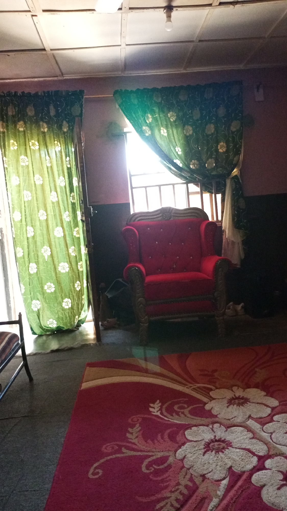 The image shows a room with a red armchair in the center. The armchair has a high back and ornate wooden trim. There is a window behind the armchair with green floral curtains. A red floral rug is in the foreground. The room appears to be in a city named Unknown, Unknown.
