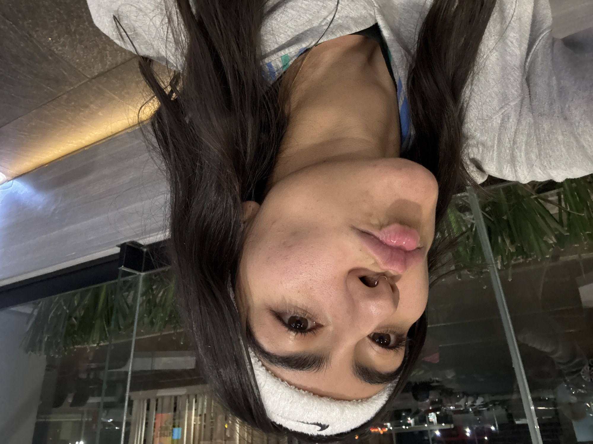 A selfie taken by a young woman with long dark hair, wearing a white Nike headband and a gray sweatshirt, possibly indoors with a reflective glass background showing a nighttime urban scene. The woman is facing the camera with slightly pursed lips. Considering the location request, it is speculative that this could be a photograph taken in Puebla, Mexico based on the prompt's additional information. The background possibly indicates an urban environment.