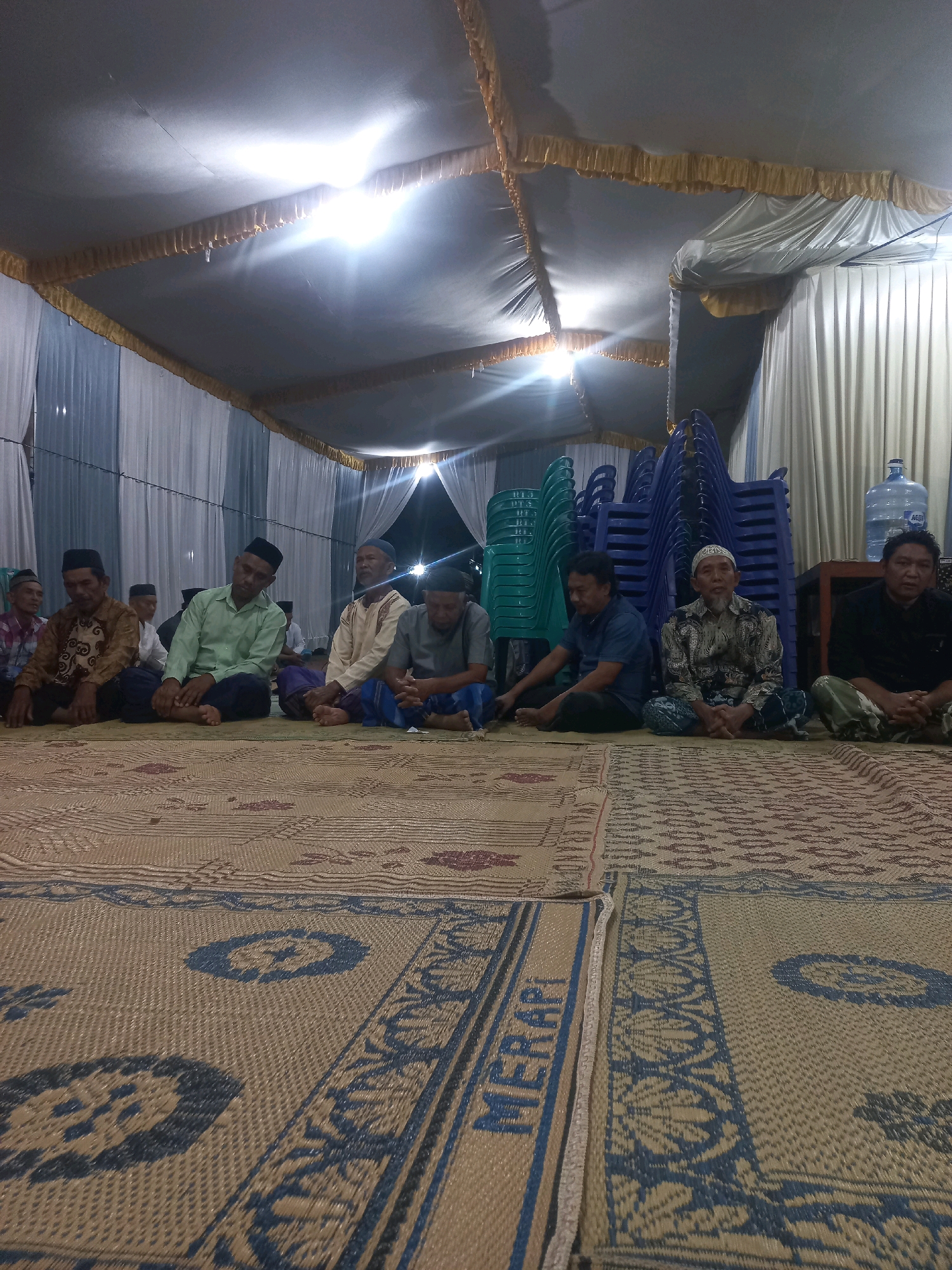 In Wiroko, Indonesia, a group of men are seated on woven mats inside a tented area. The men, dressed in traditional attire including peci caps, are positioned on the floor in rows. Some appear to be engaged in conversation, while others are looking forward or down. Behind them, stacks of blue and green plastic chairs are visible, along with a large water dispenser. The tent is illuminated by overhead lights, casting a bright glow on the scene. The environment suggests a gathering or event taking place, possibly a religious or community function.