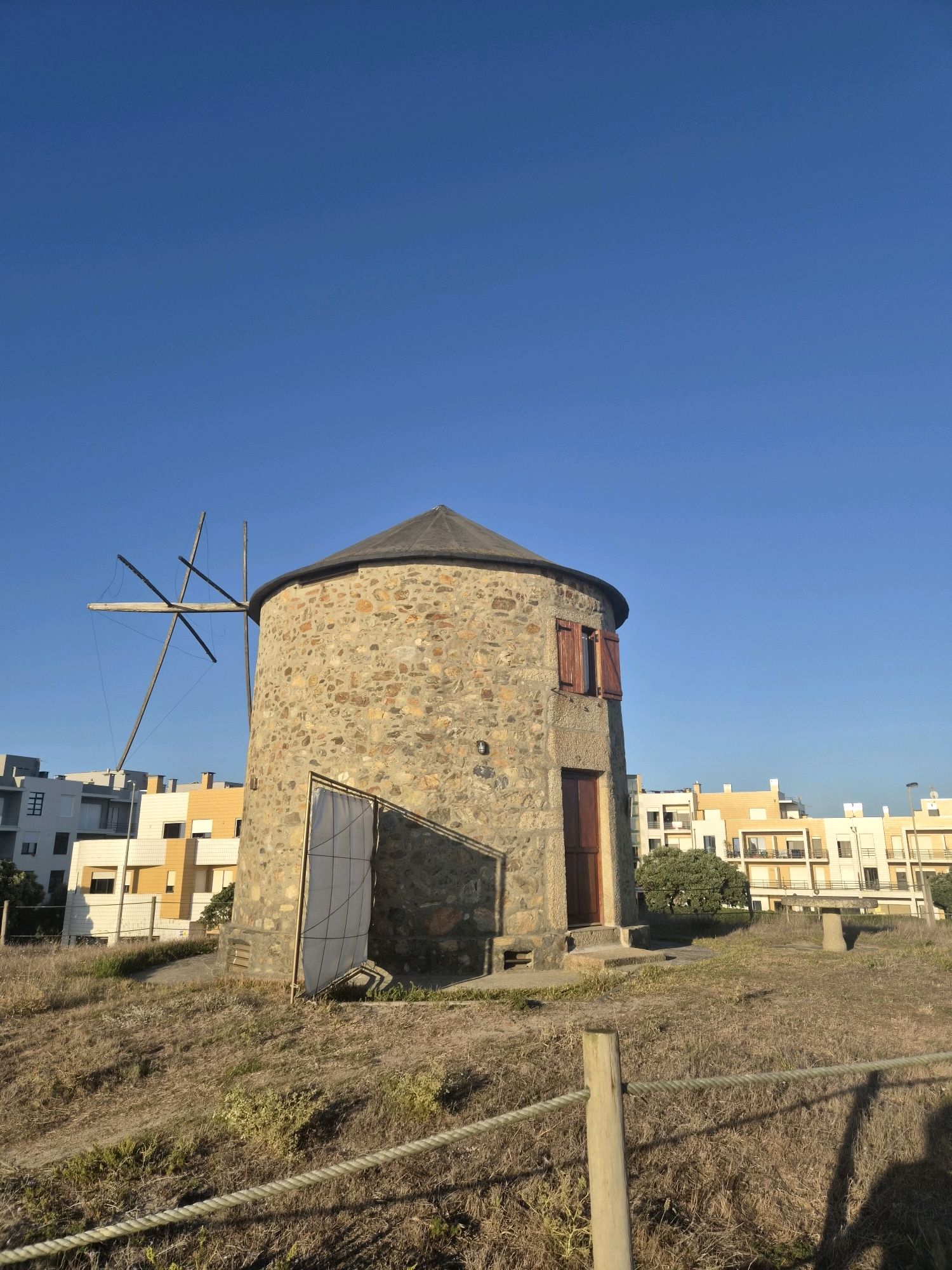 A stone windmill stands on a grassy hill under a clear blue sky. The windmill has a conical roof and two red-shuttered windows. A wooden door is visible on the side. In the background, modern residential buildings with white and yellow facades are visible. The area around the windmill is covered in dry grass and low vegetation. A rope fence runs across the foreground, with a wooden post in the lower right corner. The image appears to be taken during the daytime. The scene depicts a traditional windmill situated in a suburban or coastal area of Esposende, Portugal.