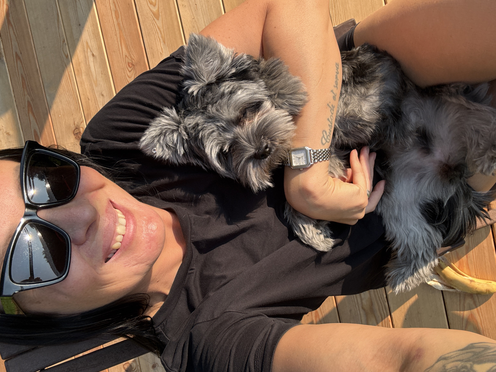 In this overhead shot, a person lies on a wooden deck with two small, gray schnauzer-type dogs. The person, wearing sunglasses and a black t-shirt, smiles widely at the camera, their head tilted back. One dog rests on the person's chest, its head nudging their arm, while the other dog lies beside them. A silver watch with a square face adorns the person's wrist. A tattoo is visible on their left arm, with the legible text "Custodi et serva." The scene is bathed in warm sunlight, suggesting it's daytime, possibly late afternoon given the long shadows cast by the deck planks. A peeled banana rests on the deck near the dogs. The setting appears to be outdoors, on a weathered wooden surface. The overall mood is relaxed and happy.