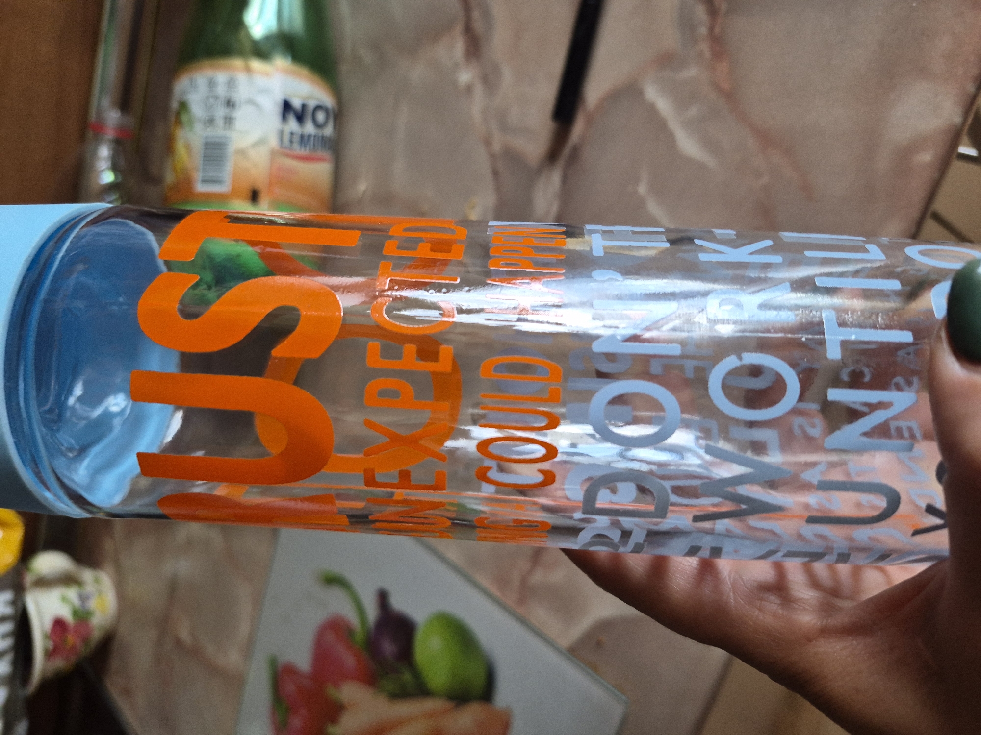 This daytime image, set in an indoor environment in Yerevan, Armenia, features a person's hand holding a clear, transparent bottle with a light blue cap. The hand, with green nail polish on the thumb, is positioned to showcase the bottle, which contains a clear liquid.
The bottle is adorned with text in two distinct colors. Prominent orange lettering spells out "JUST UNEXPECTED COULD AP," with "JUST" being the largest and most visible word. Below this, lighter blue or grey text reads "DON'T WORRY UNTIL," suggesting a motivational or reflective phrase.
In the background, a green glass bottle with a white label is visible, clearly marked "NOY LEMON." This bottle is slightly out of focus. Further details in the background include a small mug with a floral design and a cutting board or placemat featuring an image of red bell peppers and green olives. The surface beneath the items appears to be a light-colored countertop, possibly with a marble pattern. The lighting is bright and natural, indicating daylight. No specific activity is depicted beyond holding the bottle, and no emotions are discernible.