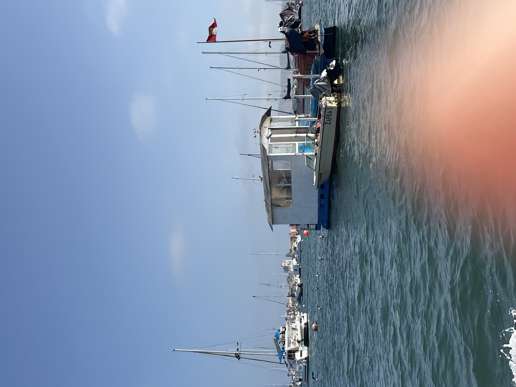 The photo shows a port in La Punta, Peru. The water is choppy and there are many boats docked. One boat is labeled "ZAFRA I". A Peruvian flag is visible on one of the boats. The sky is clear and blue.