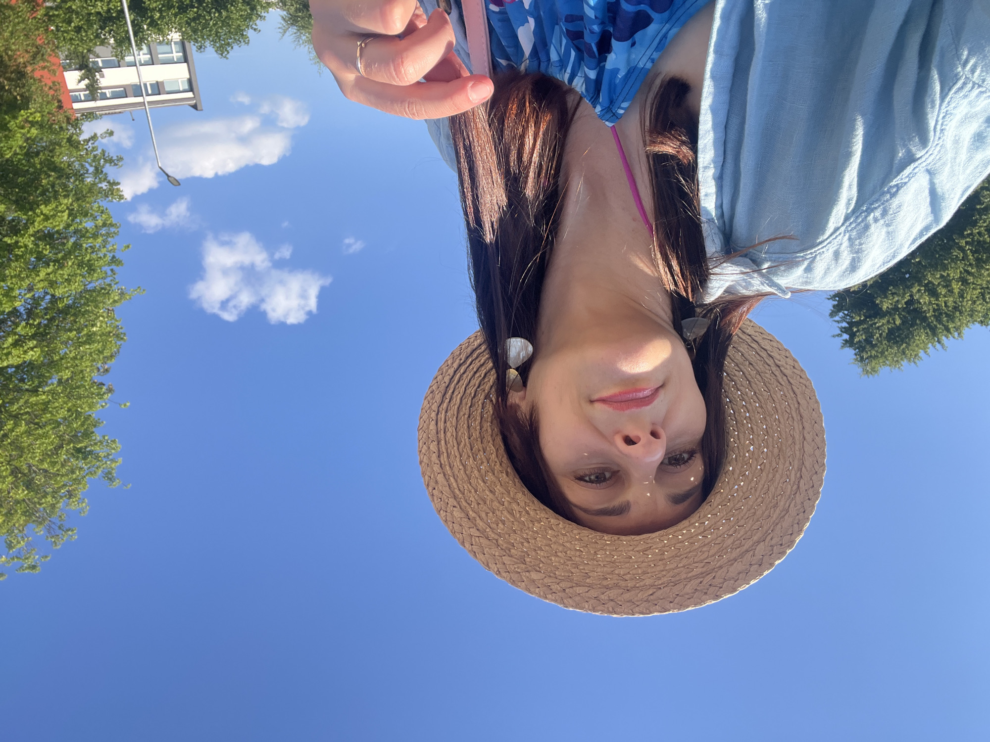 A woman with long dark hair, wearing a straw hat, a light blue collared shirt over a blue and pink patterned top, and dangling earrings, is taking a selfie outdoors under a bright blue sky with scattered white clouds. Her right hand, adorned with a ring, is visible in the foreground, near her face. In the background, trees, a street light, and a building are visible. The lighting suggests it is daytime.