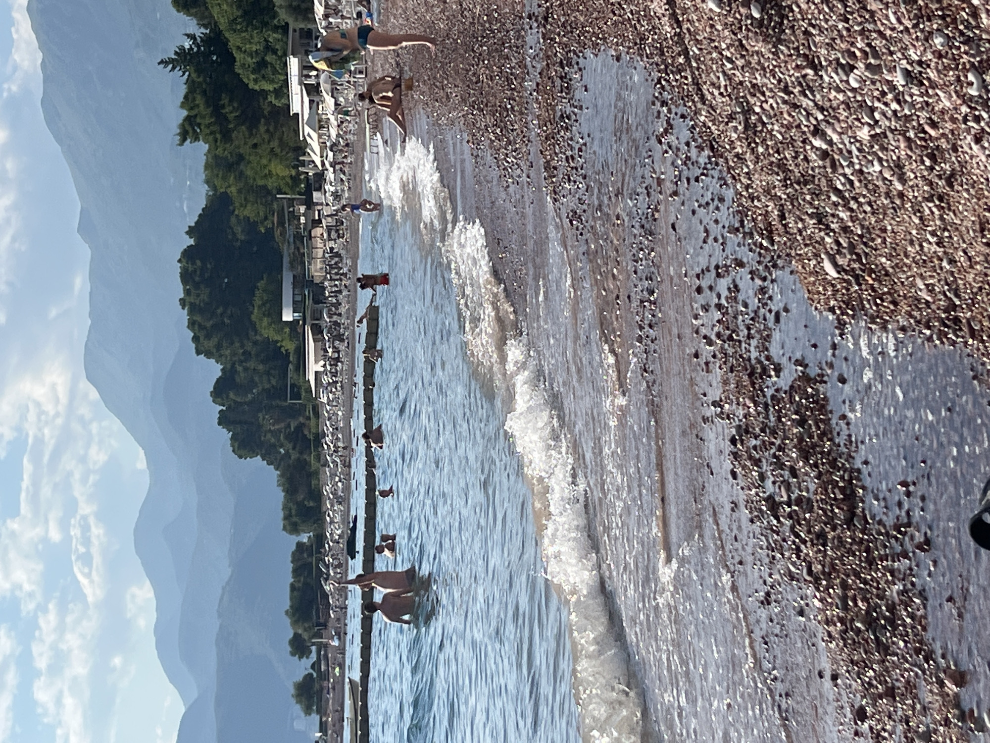 A seaside view in Sveti Stefan, Montenegro shows a pebble beach in the foreground with clear water lapping at the shore. Several people are visible in the water, some wading and others swimming. A man with his arm raised is standing in the water, facing away from the camera. Behind the beach, a row of white beach chairs and umbrellas stretches along the coastline. Lush green trees and the hazy silhouettes of mountains form the backdrop under a partially cloudy sky.