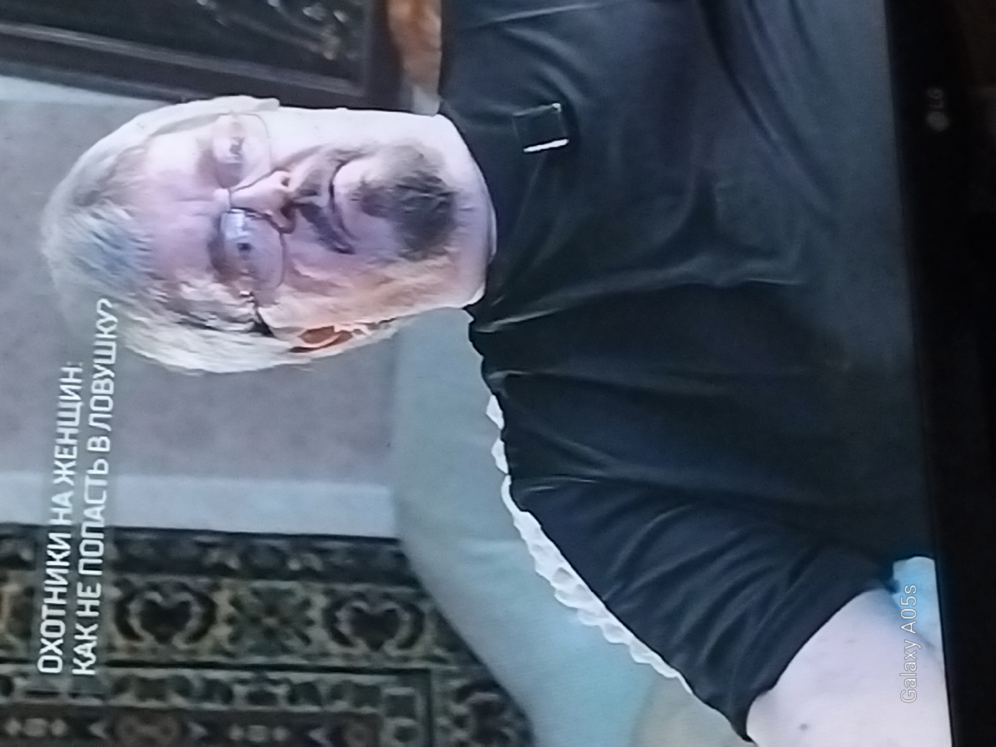 The image shows a man wearing glasses and a black t-shirt with a small black clip attached to the collar area. He has graying hair and a mustache and goatee. The text superimposed on the image reads "ОХОТНИКИ НА ЖЕНЩИН КАК НЕ ПОПАСТЬ В ЛОВУШКУ?". This translates to "Hunters for Women: How Not to Fall into the Trap?". The background shows a patterned rug and a light-colored sofa or chair. The bottom left corner displays the text "Galaxy A05s", indicating the device used to capture the image, and the bottom right corner shows the LG logo. The location is identified as Druzhba, Russia. The scene appears to be an indoor setting, possibly a television broadcast or a personal recording.