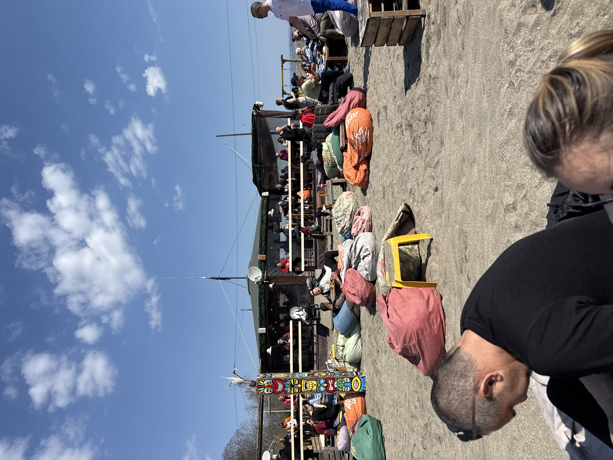 The scene captures an outdoor establishment, potentially a beachside cafe or bar, situated in Burgas, Bulgaria, evident from the urban setting and beach access. The area is populated with individuals enjoying the atmosphere; some are seated at elevated tables while others relax on beanbag chairs spread across the sandy ground. A colorful totem pole adds a unique visual element, standing alongside wooden structures and assorted seating arrangements. The sky is clear with scattered clouds, indicating a sunny day. Several overhead power lines stretch across the top of the frame. A mix of individuals is captured in the foreground, including a person with a close-cropped haircut wearing sunglasses, and another with blonde hair partially visible. The overall impression is that of a relaxed outdoor social space, likely popular during warmer weather.
