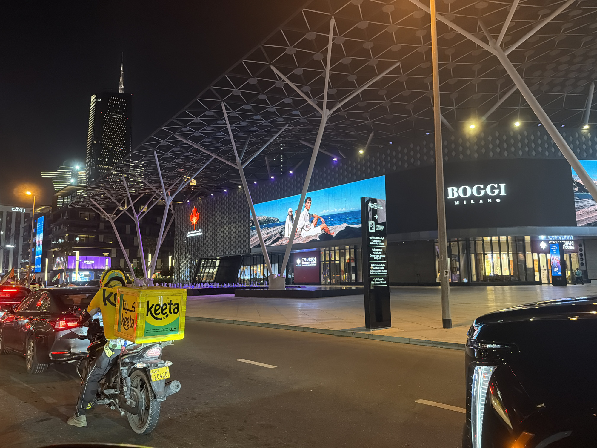 This image captures a vibrant, artificially illuminated urban street scene at night in Dubai, United Arab Emirates, specifically in the City Walk area, with the Burj Khalifa discernible in the distant background.
In the foreground, on the right, the lower section of a black vehicle, likely an SUV, is visible, with its bright white vertical daytime running light illuminated. To the left, a motorcycle delivery rider is stationary, facing away from the viewer. The rider is dressed in a yellow long-sleeved shirt and helmet, both emblazoned with the "keeta" logo. A bright yellow cubic delivery box, also branded with "keeta" in both Latin and Arabic script, is mounted on the rear of the black motorcycle. The motorcycle's rear license plate displays "DUBAI 20430". Directly behind the motorcycle, a dark-colored sedan with illuminated taillights is also stationary, indicating traffic.
The paved street in the mid-ground features a single white broken lane marker. To the right of the street, a wide, clear pedestrian walkway extends towards a complex of modern buildings. A tall, slender black signpost stands upright on the walkway, partially obscuring text. Overhead, a distinctive white, angular, tree-like lattice structure forms a canopy, providing architectural interest.
The background is dominated by contemporary multi-story buildings adorned with large, illuminated digital displays. A prominent screen on the right showcases "BOGGI MILANO" in white text against a black background. To its left, another expansive screen displays an advertisement featuring two men in white attire by a body of water. Further left, a building facade shows a red maple leaf symbol and the text "CANADIAN," likely belonging to Canadian University Dubai, which has a presence in the area. Other structures with various light patterns and a fountain feature with purple illumination are also visible. In the far background, the iconic, tall silhouette of the Burj Khalifa stands prominently, its upper sections and spire brightly lit against the dark night sky. The entire scene is bathed in artificial light from street lamps, building lights, and digital signage, confirming the nighttime setting.