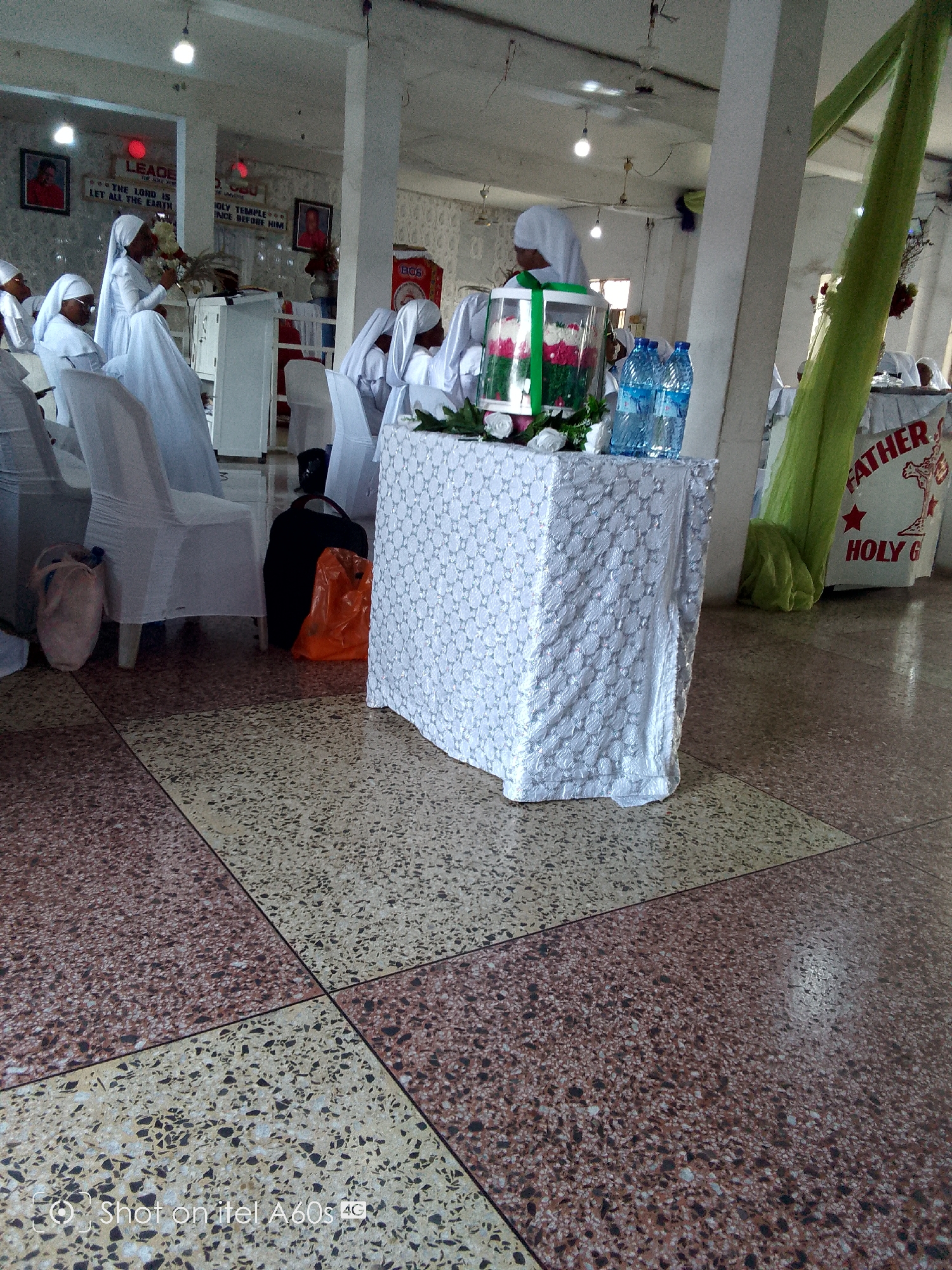 The image captures a scene within a building in Lagos, Nigeria, identifiable by the English language signage, the style of dress of the people, and the "Shot on itel A60s 4G" watermark from a mobile phone likely used in the region. The interior appears to be a place of worship or a community hall, as suggested by the individuals dressed in white gowns and head coverings, typically associated with religious ceremonies or gatherings.
Several people are present, some seated on white-covered chairs, seemingly observing or participating in an event. A white-covered table prominently displays decorative items, including flowers in a clear container with a green ribbon, and bottles of water. Behind the table, a banner with the words "FATHER" and "HOLY G" is visible, further implying a religious context.
Bags are placed under and next to the chairs and table, indicating personal belongings. The floor is tiled with a speckled pattern.