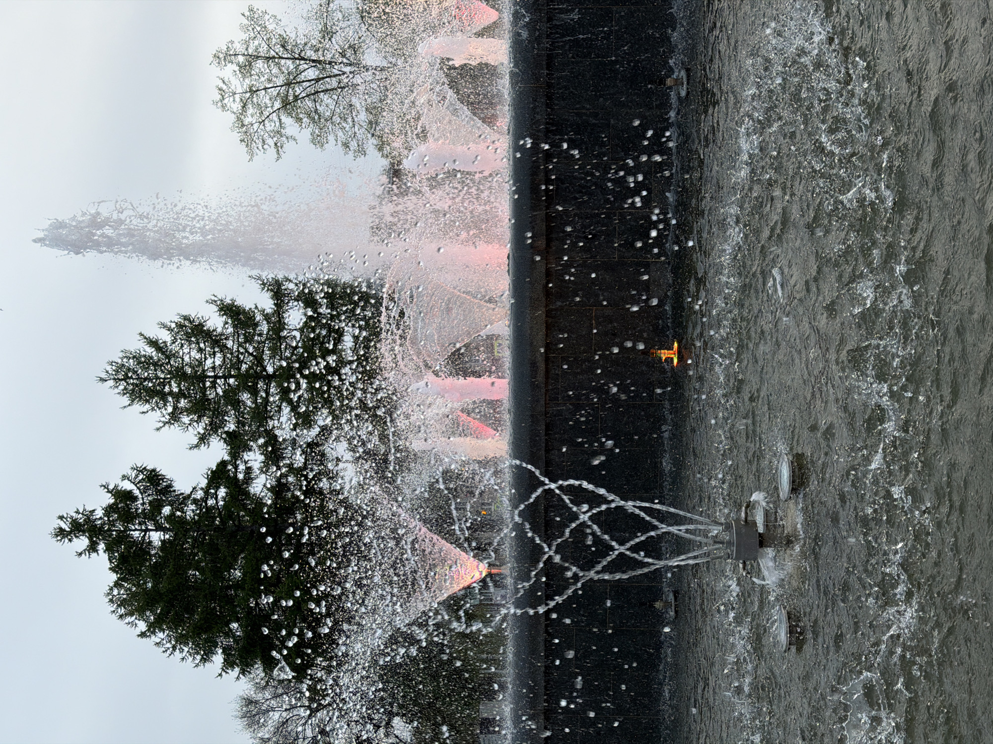 This image shows a fountain in the city of Chaikovsky, Russia. The fountain has multiple levels with water spraying from various points. Some of the water jets are illuminated with pink or orange lights. The fountain is constructed with dark, tiled surfaces and is filled with water. Trees are visible in the background, suggesting the fountain is located in a park or public area. The sky is overcast.