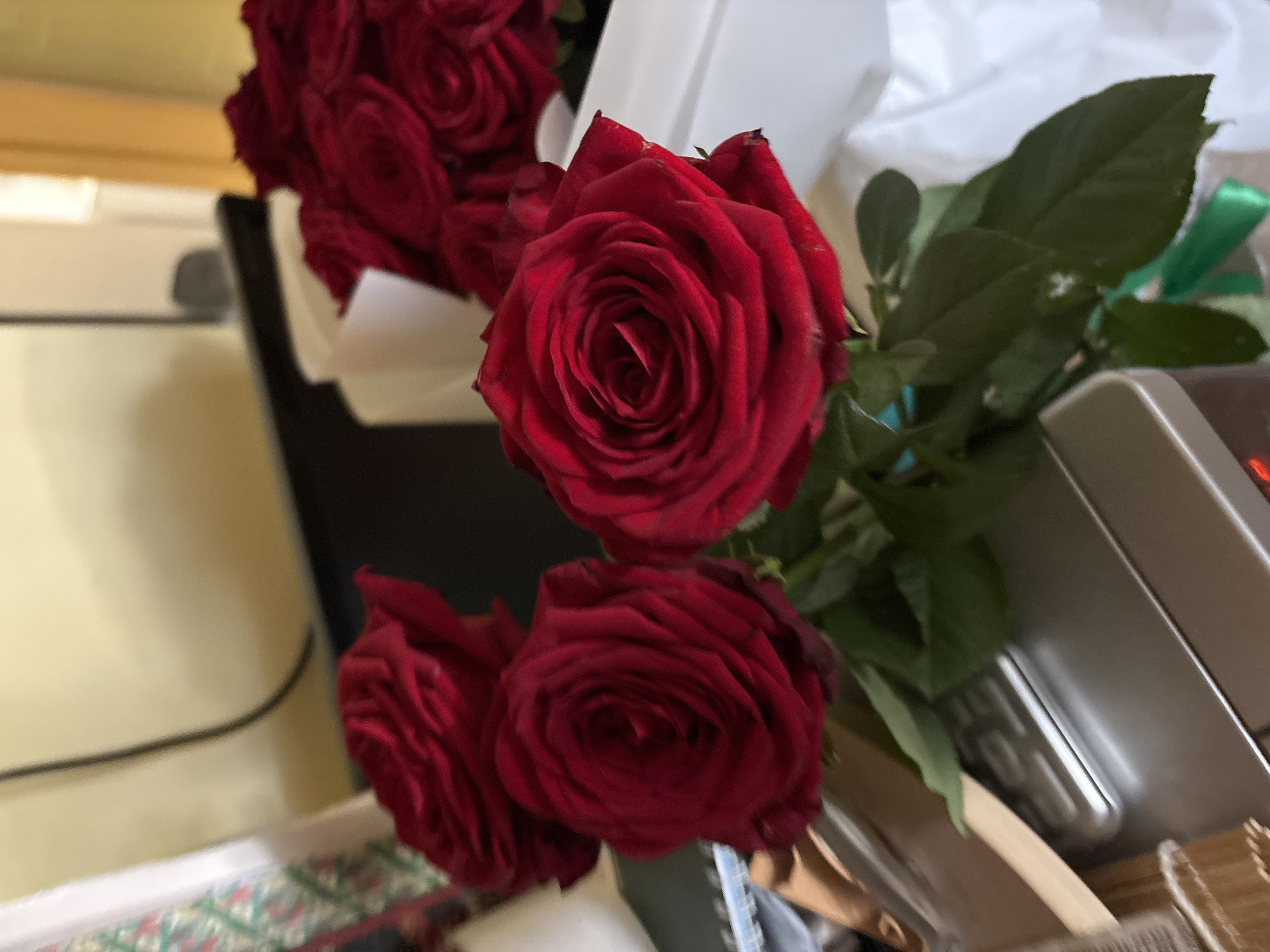 A close-up, slightly blurry photograph shows a bouquet of red roses. The roses are deep red with velvety petals and are arranged with green leaves. Some of the roses are in focus in the foreground, while others are out of focus in the background. The roses appear to be in a vase or container, partially obscured by white wrapping and a green ribbon. In the lower part of the frame, there is a silver-colored electronic device, possibly a scale, with some wooden objects and a white patterned item to the left. The background is indistinct, with a light-colored wall and a dark object. The lighting is warm and appears to be from an artificial source. The image does not contain any recognizable landmarks or contextual information that would indicate its location in Minsk, Belarus.