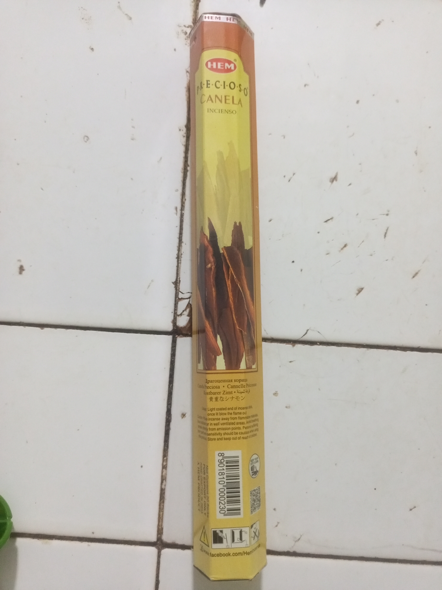 The image shows a box of HEM brand incense sticks with a cinnamon scent. The box is predominantly yellow with an illustration of cinnamon sticks. Text on the box is in multiple languages, including Russian, Spanish, French, German, Arabic, and Japanese, as well as English. The English text includes "PRECIOUS CINNAMON INCENSE" and "Caution: Place incense away from flammable materials. Burn in well ventilated areas. Avoid breathing smoke. Persons suffering from respiratory sensitivity should be cautious when using the product. Store and keep out of reach of children." A barcode and a QR code are also visible on the box. The box is positioned vertically on a white tiled surface. The tiles are grimy, with dark grout lines and visible dirt and debris, particularly in the grout. The lighting appears to be artificial and somewhat dim. There is no indication of the location being La Tagua, Venezuela.