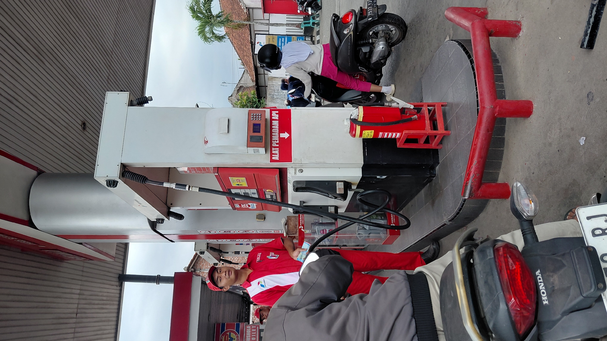 The image depicts a gas station in Rejasari, Indonesia. A gas station attendant in a red and white uniform is standing next to a gas pump, holding money. In the foreground, a person on a motorcycle is getting gas. Another person on a motorcycle, wearing a helmet and pink pants, is also at the gas station. A fire extinguisher is visible on a red stand. The environment appears to be an outdoor setting with a covered canopy and a cloudy sky. Buildings and palm trees are visible in the background.