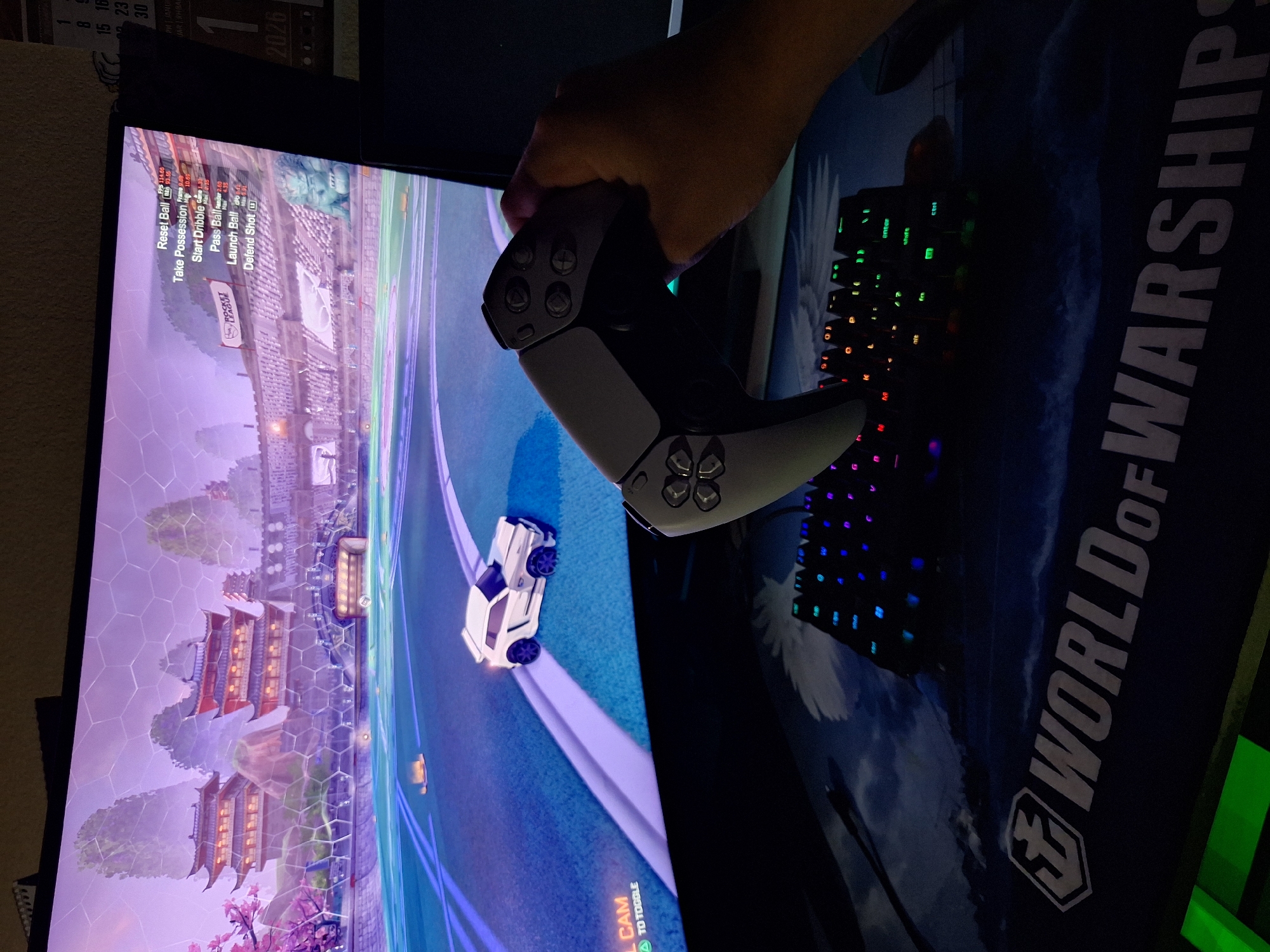 A first-person perspective shot captures a hand holding a white PlayStation 5 controller in front of a large computer monitor. The monitor displays a scene from the video game Rocket League, featuring a futuristic cityscape with Asian-inspired architecture and a digital soccer field. The controller is positioned in the lower right quadrant of the image, partially obscuring the screen. Below the monitor, a black keyboard with illuminated keys rests on a dark blue mousepad. The mousepad has a graphic of a ship and the text "WORLD OF WARSHIPS" printed on it. The background is dark, suggesting the image was taken in a dimly lit room. The scene is set in an urban environment.