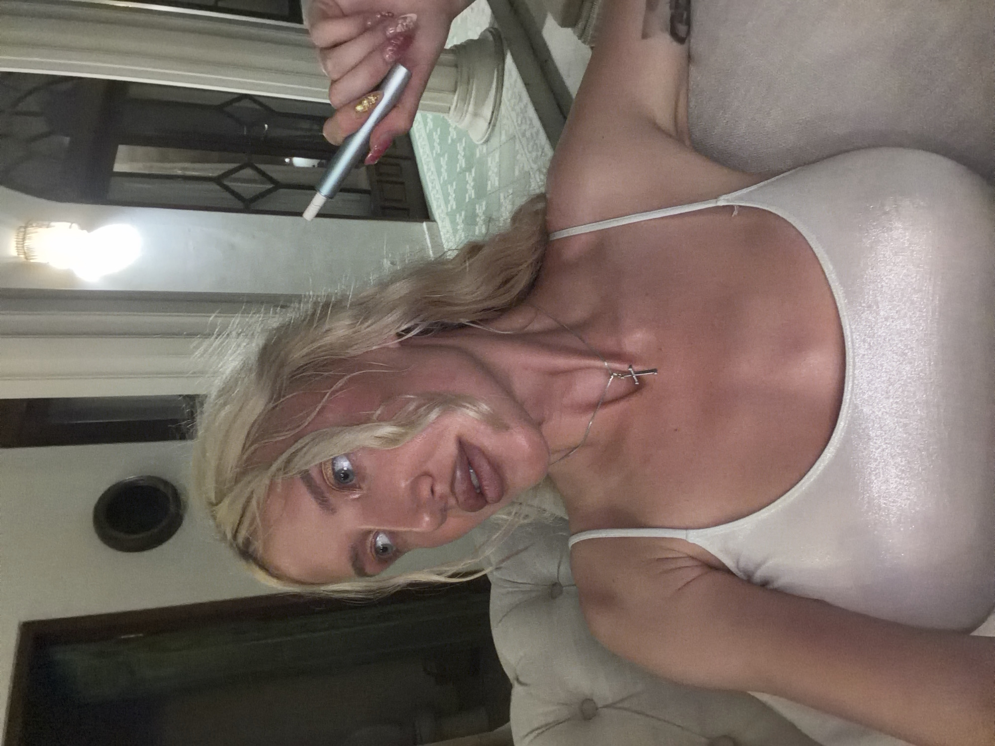 A blonde woman sits on a plush white sofa in what appears to be a hotel lobby or high-end establishment, possibly in North Kuta, Indonesia. The woman, wearing a sparkly white camisole, holds a silver e-cigarette in her right hand. Her makeup is prominent, with dark eyeliner and lip gloss. A silver cross necklace adorns her neck. The background shows ornate architecture, including large pillars, patterned flooring, and an arched doorway leading to another room. A light fixture hangs from the ceiling. The lighting is dim, suggesting it could be evening. The overall impression is one of casual luxury and relaxation.
