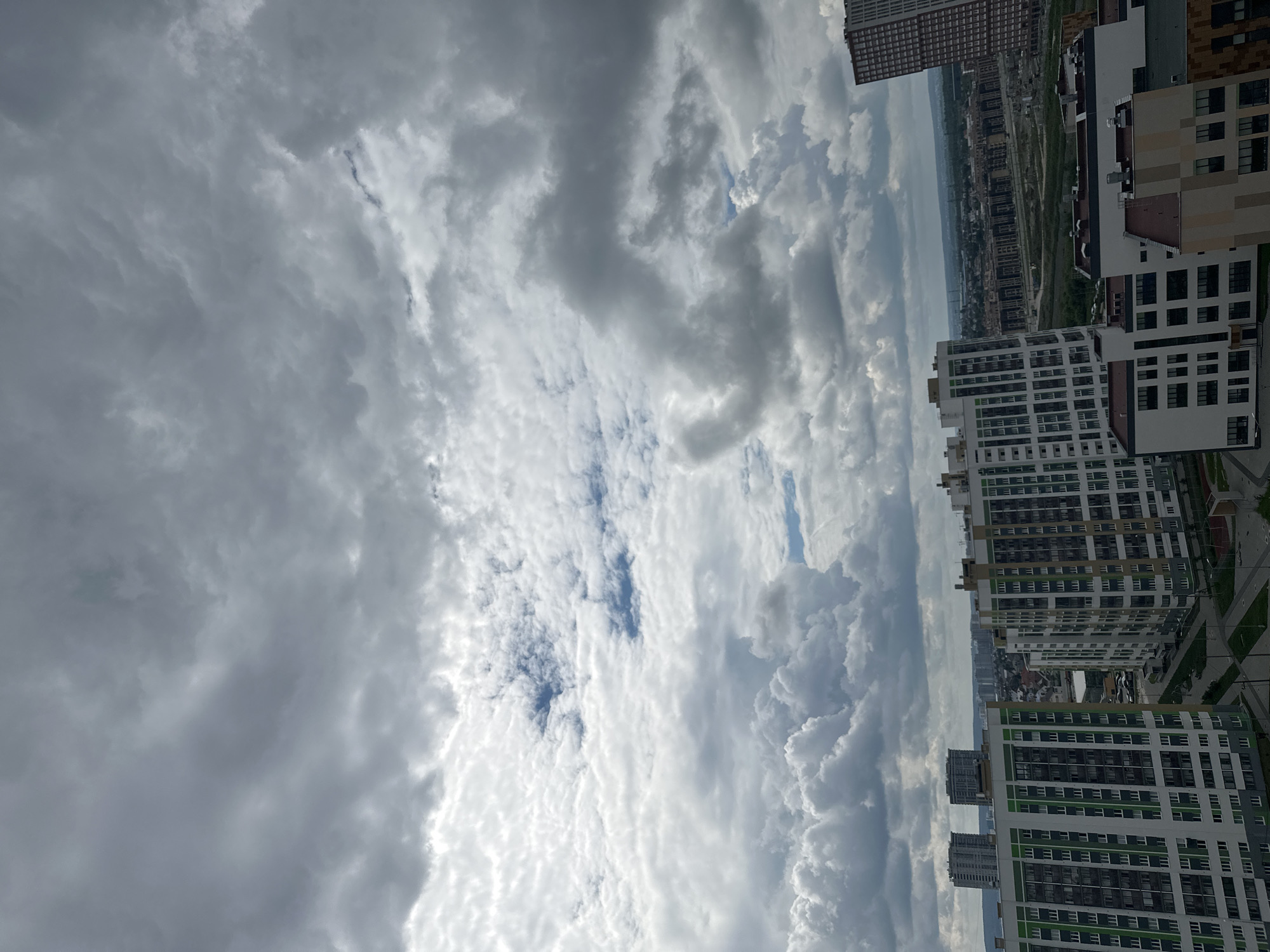 The image presents an elevated view of a modern urban landscape in the Novokoltsovsky microdistrict of Yekaterinburg, Russia. The sky occupies the upper two-thirds of the frame, dominated by a dense, varied cloud cover featuring large, white cumulus clouds intermingled with expansive gray stratiform clouds, with intermittent small patches of blue sky visible, suggesting a partly cloudy to overcast daytime.
In the foreground and midground, several newly constructed, multi-story residential buildings are prominent. On the left, a large, contemporary building features a white facade accented with prominent vertical sections of green and dark gray panels, perforated by numerous windows and balconies. To its right, another similar-style building is visible, distinguished by white, beige, and dark gray facade elements. Further back on the left, two taller, darker high-rises stand in the distance. The lower right foreground includes additional shorter residential or auxiliary structures with varied beige, brown, and dark gray cladding. The ground level immediately surrounding these buildings shows planned urban landscaping with paved areas and green spaces.
The background extends into a broad urban sprawl, characteristic of Yekaterinburg, with a multitude of buildings of varying heights and ages stretching towards a distant, hazy horizon. No individuals or specific activities are discernible within the frame.