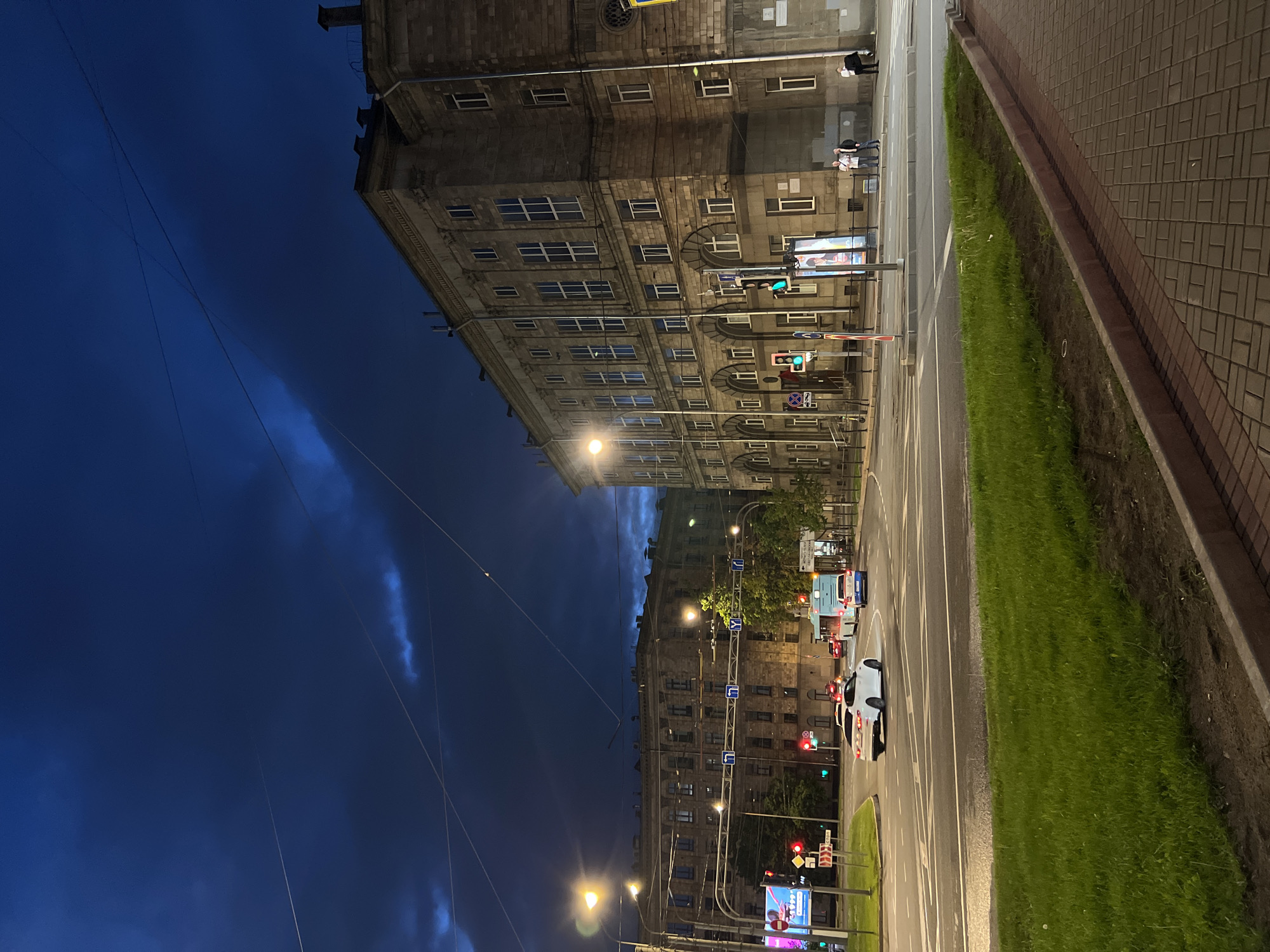 The image depicts a street scene at dusk in an urban environment, possibly in a city like St. Petersburg, Russia, given the architectural style and signage. A large, multi-story stone building with numerous windows dominates the right side of the frame, featuring classical architectural elements. Across the street, another similar building is visible in the background. The sky is a deep blue, with some dark clouds and thin wisps of lighter clouds, suggesting an overcast or post-storm condition. There are overhead power lines crisscrossing the sky.
The foreground shows a paved sidewalk and a grassy median strip. The street is a multi-lane intersection with visible lane markings. Several vehicles are present, including a white sedan turning at the intersection, a blue and white bus, and other cars in the distance. Traffic lights are illuminated with red and green signals. Pedestrians can be seen on the sidewalk near the building on the right, with two individuals standing together and another two further down. A streetlamp is lit on the left side of the image, casting a warm glow. Advertising displays are also present on the left and right sides of the intersection. The overall impression is a busy urban intersection at the transition from day to night.