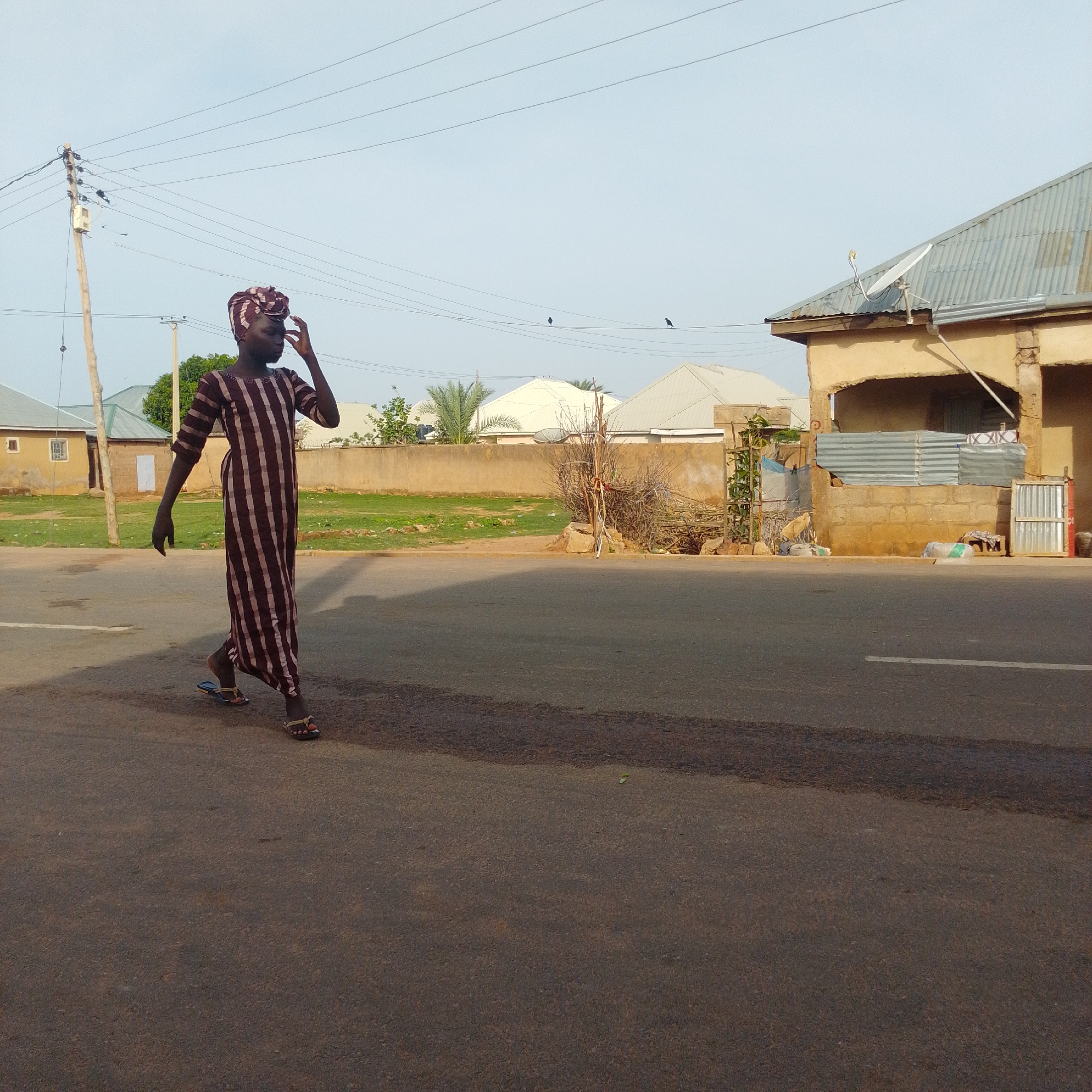 The image depicts a child wearing a long, striped garment and a headscarf, walking on a paved road in Gombe, Nigeria. The child is on the left side of the frame, mid-stride, with their right hand touching their forehead. In the background, there are residential buildings with corrugated metal roofs, a concrete wall, some vegetation including palm trees, and utility poles with overhead wires. The sky is clear and pale. The road surface appears to be asphalt, with a white line visible on the left side. The scene appears to be in a residential area of Gombe, characterized by the typical architecture and infrastructure seen in the region.