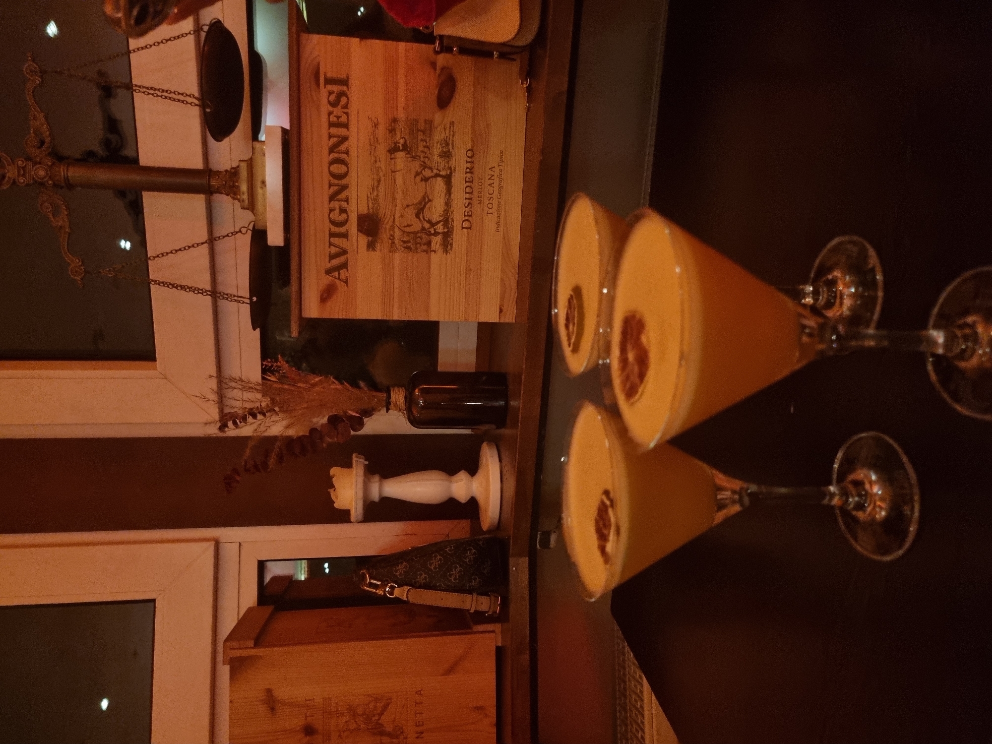 On a dark brown table in a dimly lit location in Saarbrücken, Germany, two cocktail glasses, filled with an orange-colored beverage and garnished with a dark topping, are reflected in the surface. Behind the table, an arrangement includes an antique balance scale, a wooden box labeled "AVIGNONESI" and "DESIDERIO TOSCANA," a small decorative glass bottle, a white candlestick with a partially melted candle, and a handbag with a patterned design. The scene is composed near a window with a white frame.